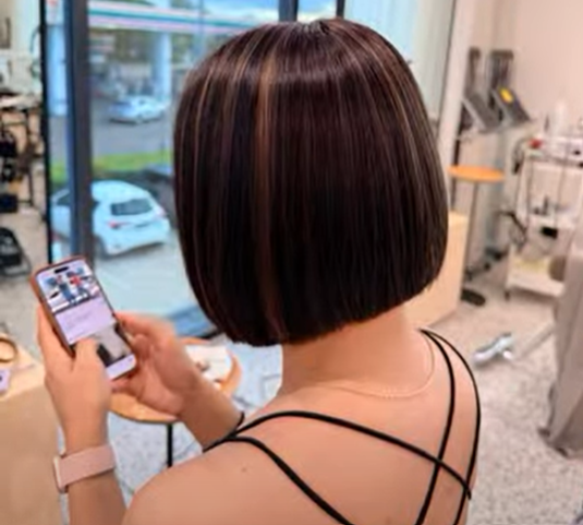 Back view of a woman with a short, sleek, dark brown bob haircut, wearing a black strappy top, looking at her smartphone inside a salon.