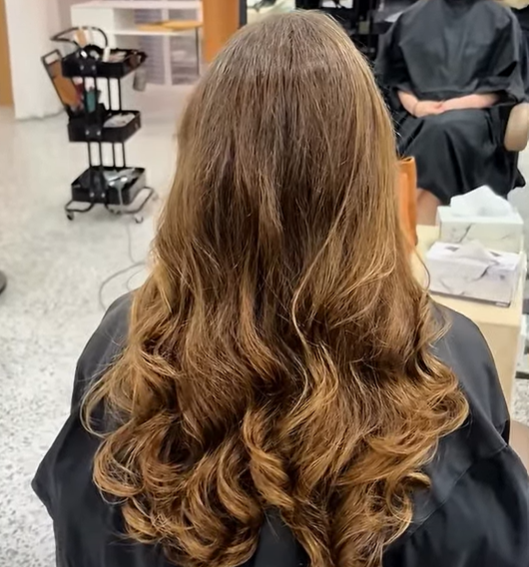 Back view of a woman with long, wavy, light brown hair sitting in a salon chair.