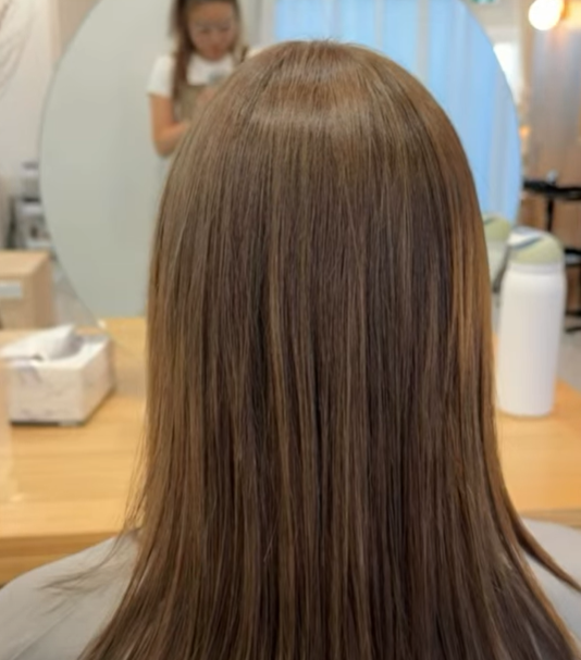 Back of a person with straight, shiny, light brown hair in a salon, with a child in the background doing a craft.