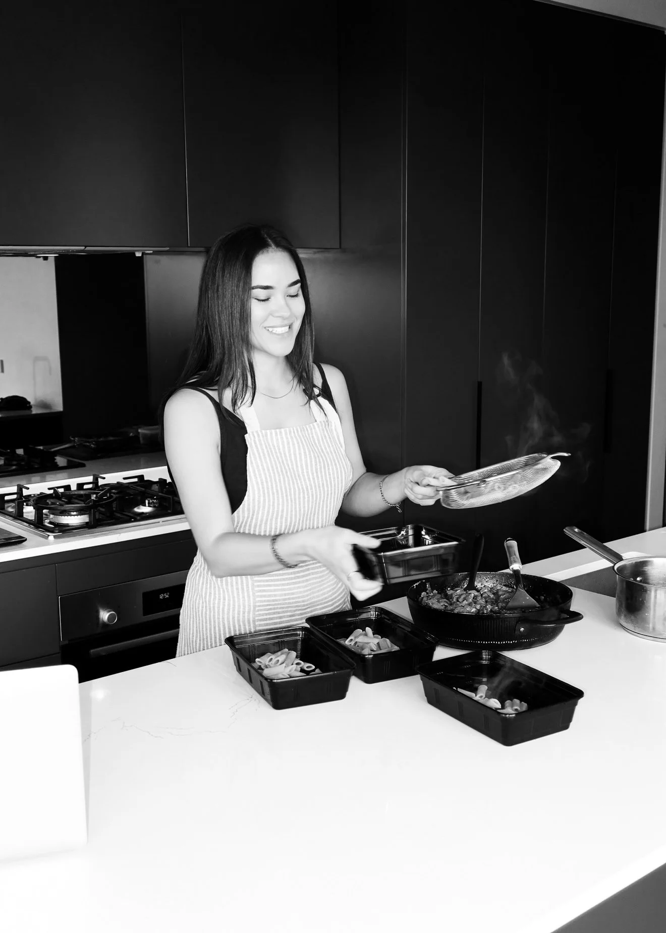 A woman cooking in a modern kitchen, wearing an apron and smiling, with food containers on the kitchen counter.