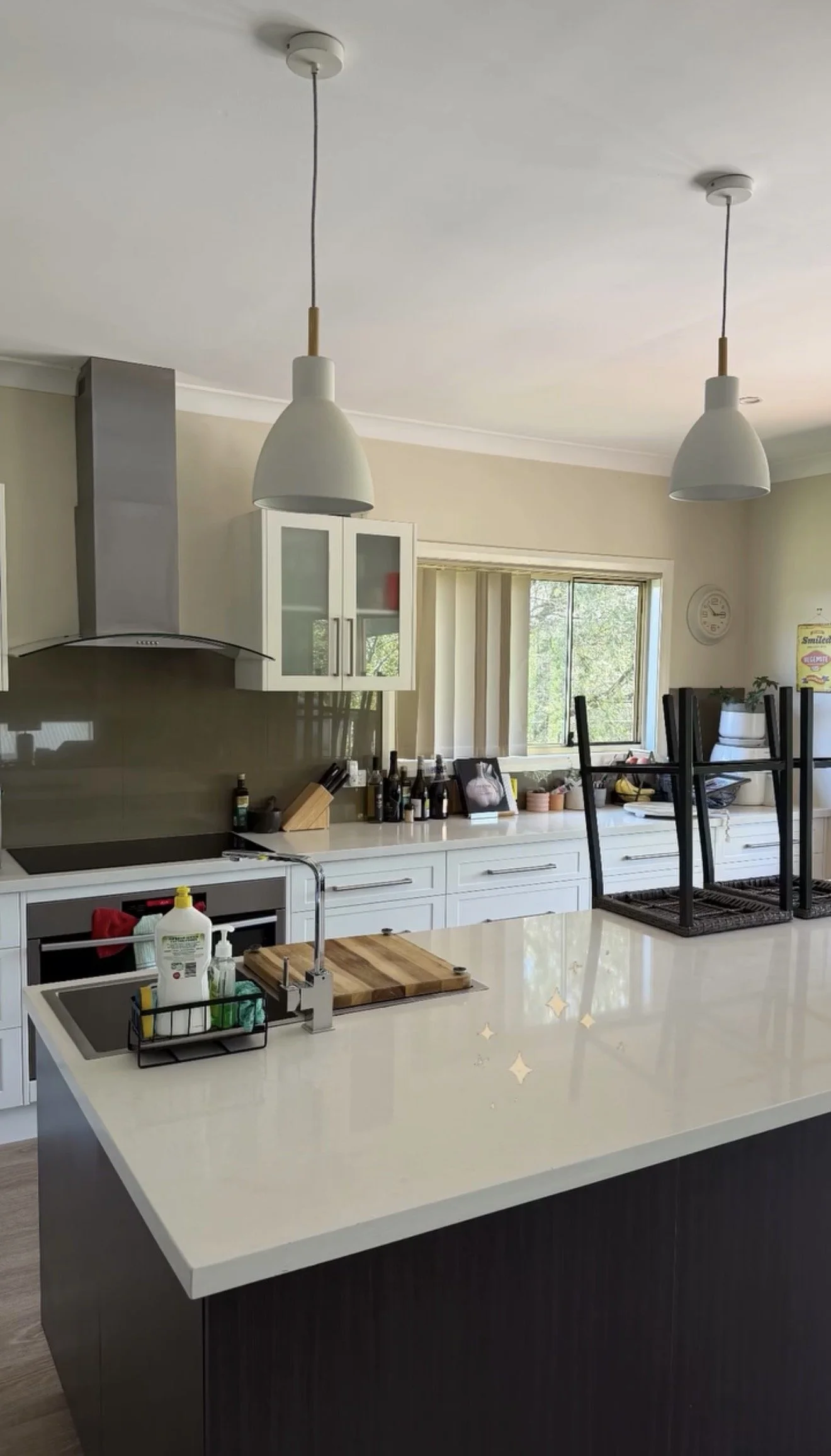 Modern kitchen with white countertop, hanging lights, a window, and various items on the counter, including bottles, framed photo, and decorative objects; chairs are stacked upside down on the island.