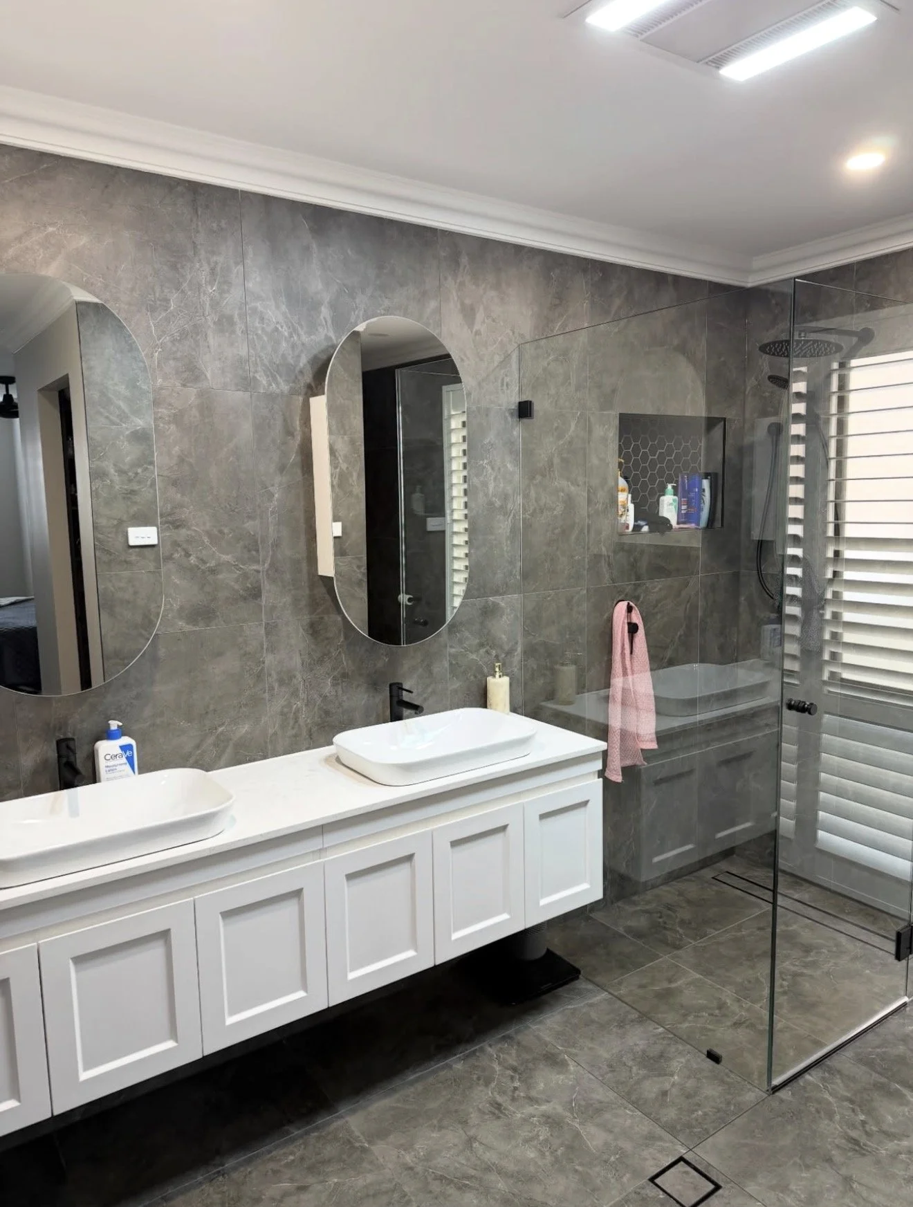 Modern bathroom with gray marble tiles, a white double vanity with vessel sinks, and a glass-enclosed shower area with a rainfall showerhead. There are two mirrors above the sinks and a pink towel hanging on a hook.