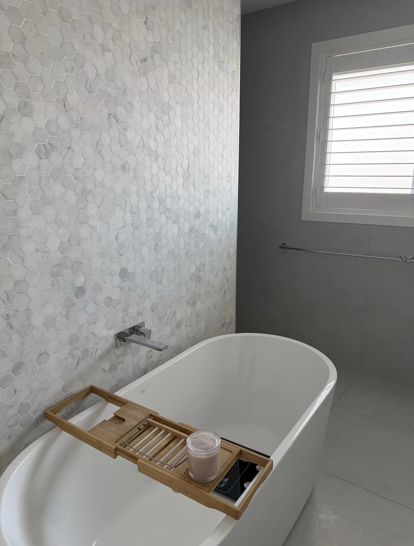 Bathroom with white hexagonal tiled wall, a freestanding bathtub with a wooden bath tray holding a candle and a glass of water, gray walls, and a window with white shutters.