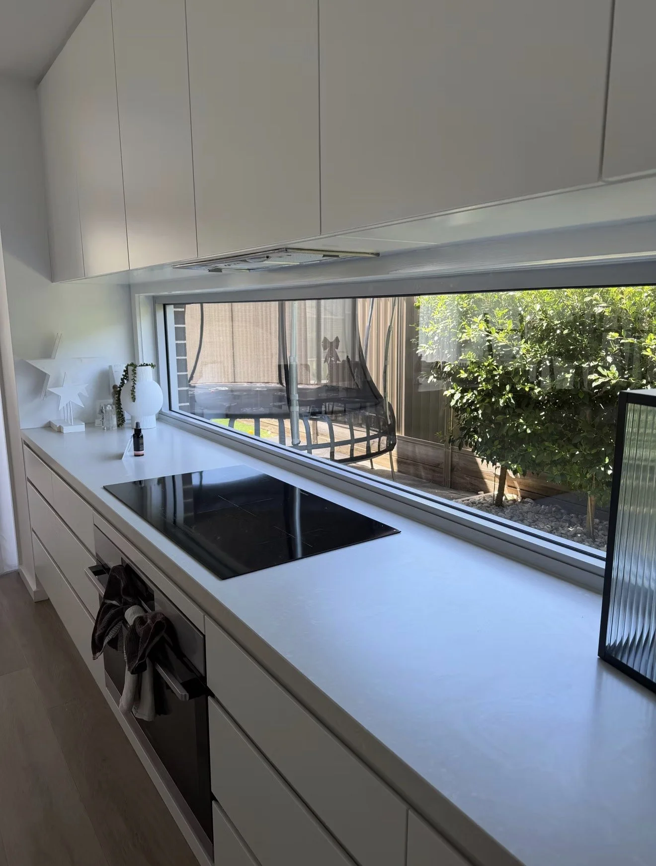 Modern kitchen with white cabinets and countertop, black cooktop, decorative items, and large window overlooking a backyard with greenery, trampoline, and patio.