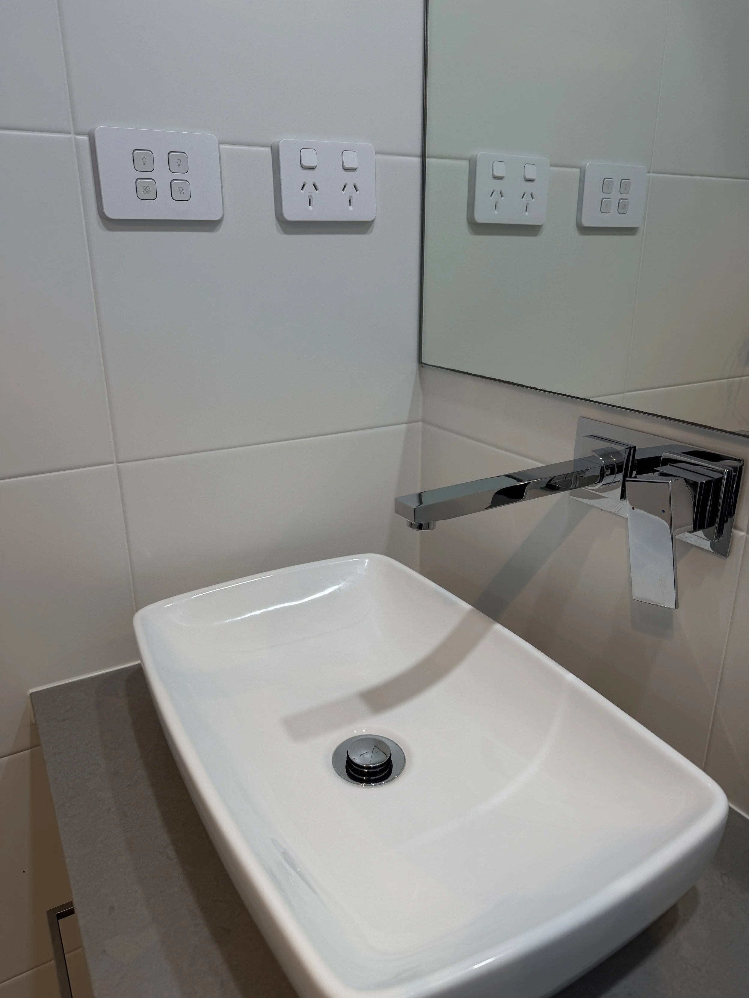 A white bathroom sink with a chrome faucet and a circular drain. Behind the sink, there are three electrical outlets and switches on the tiled wall, with a mirror reflecting the outlets. The countertop is made of gray material.