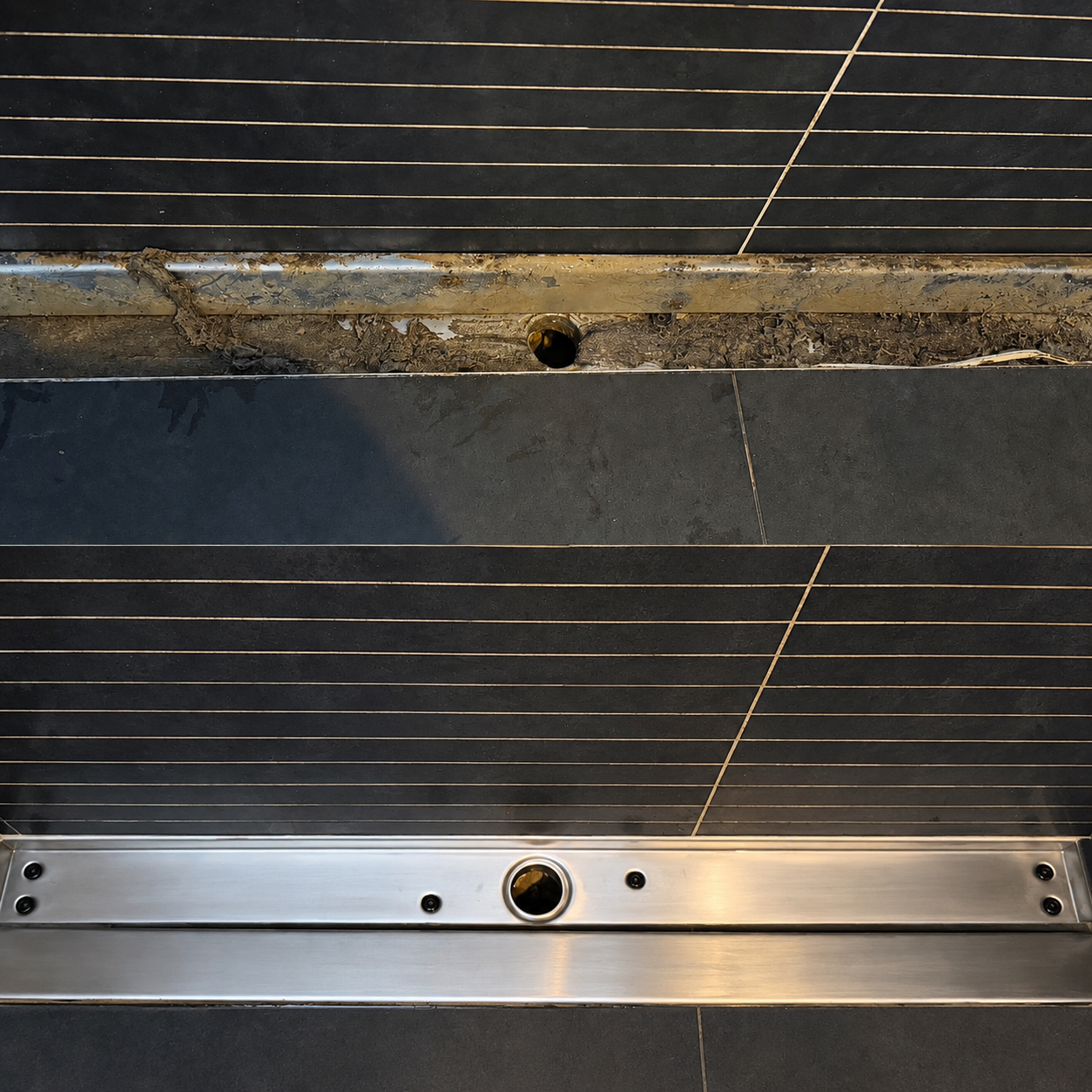 Close-up of a tiled bathroom or shower area with missing or removed fixtures, showing a hole in the wall with dirt and debris, and an empty drip tray or drainage area.