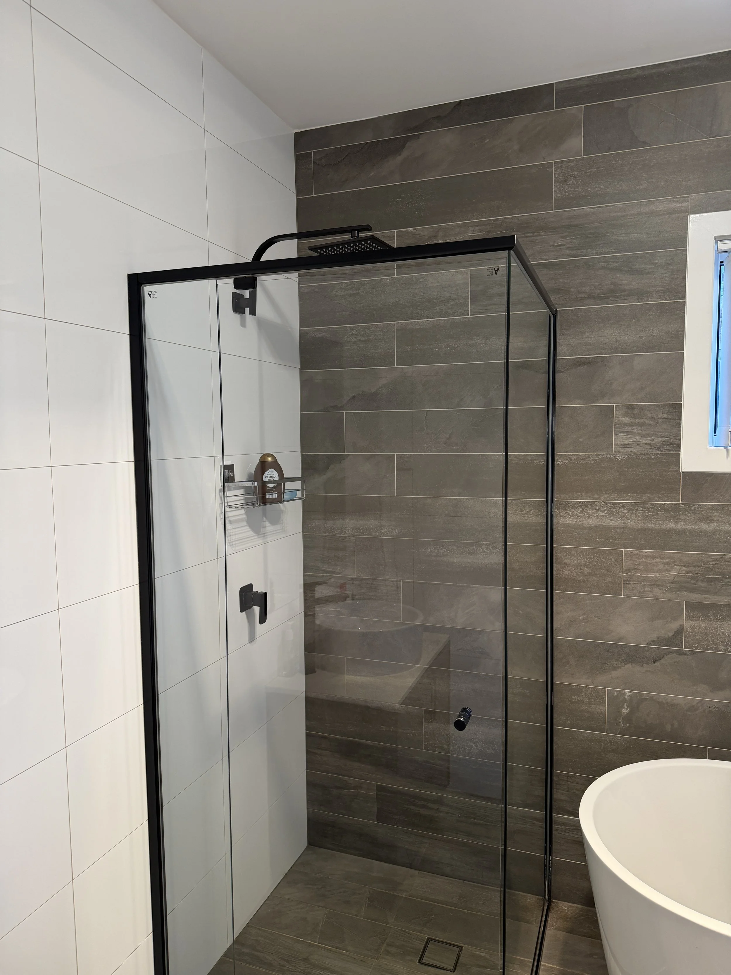 Modern bathroom with a walk-in shower featuring a black frame, large gray and white tiles, a small window, and a freestanding bathtub.