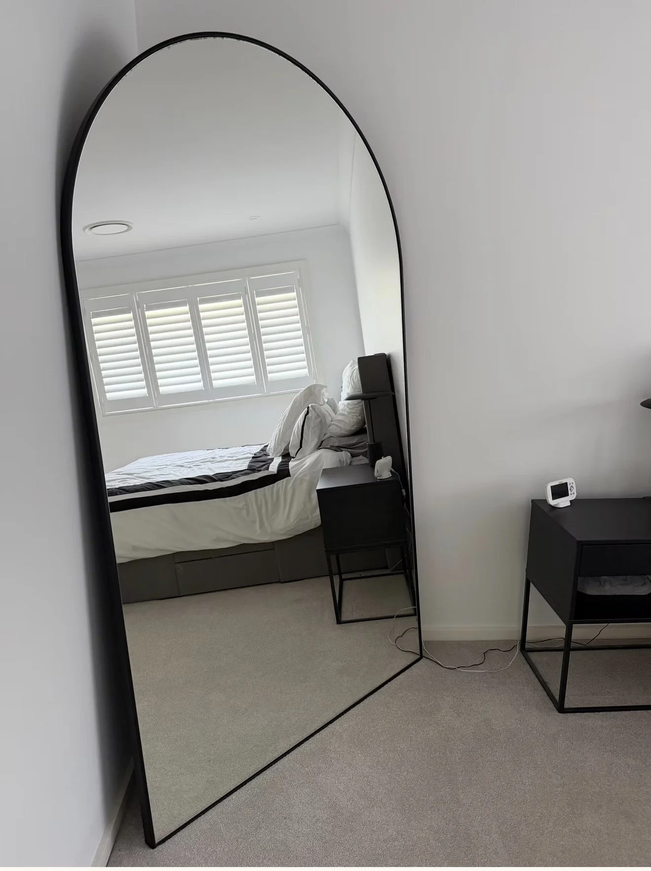 A bedroom reflected in a full-length mirror, showing a bed with white and black bedding, a window with white blinds, a small black table with a clock, and a black dresser on the wall.