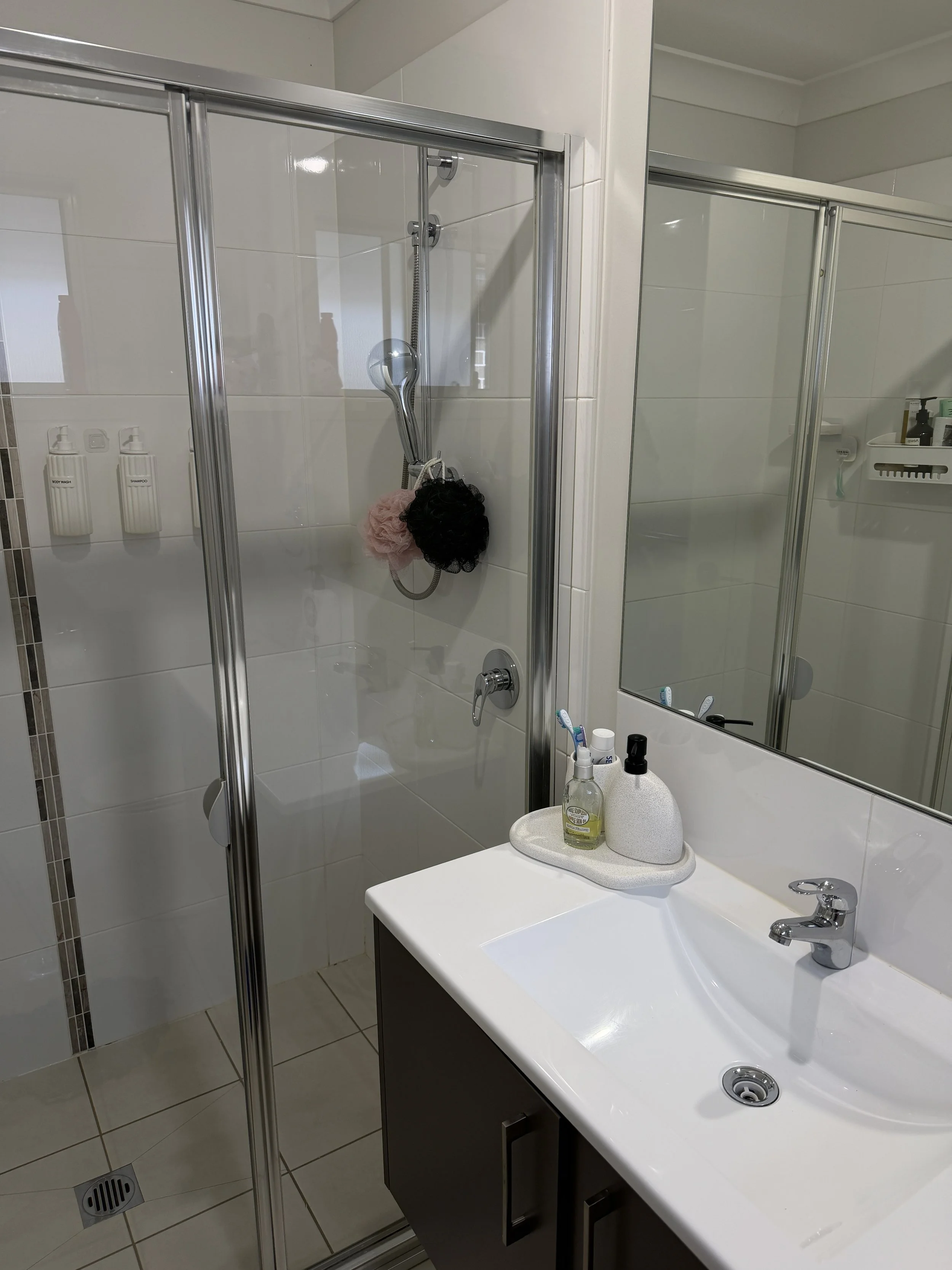 Bathroom with a shower stall, white tiled walls, and a white sink. The shower has a glass door, a silver shower head with a hose, and soap dispensers labeled 'SOAP WASH' and 'SHAMPOO'. There is a mirror above the sink with toothbrushes and bottles on the counter.