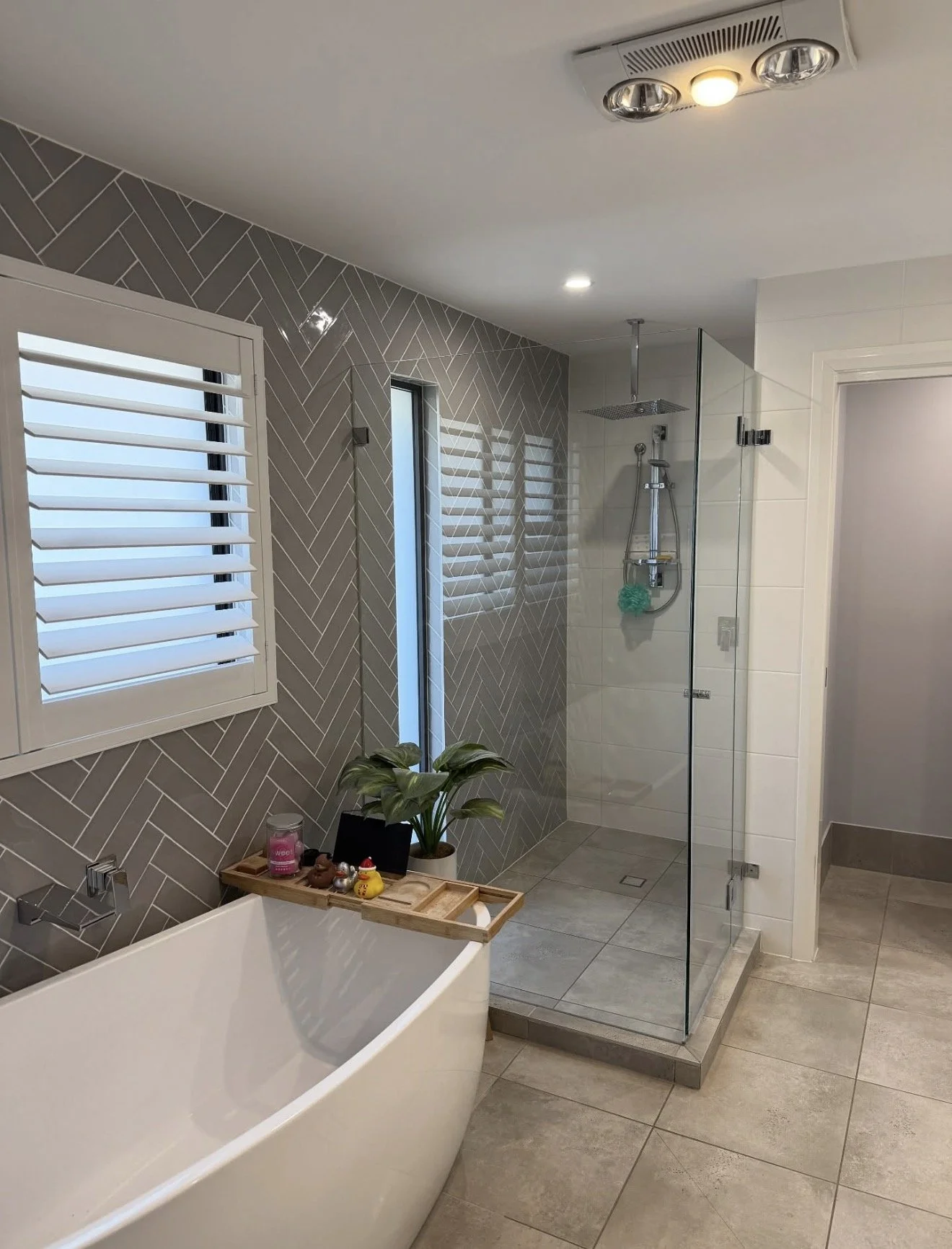 A modern bathroom with a white bathtub, a gray tile wall, a small wooden tray with various small items on the edge of the bathtub, a potted plant, a shower with glass enclosure, and a white window with blinds.