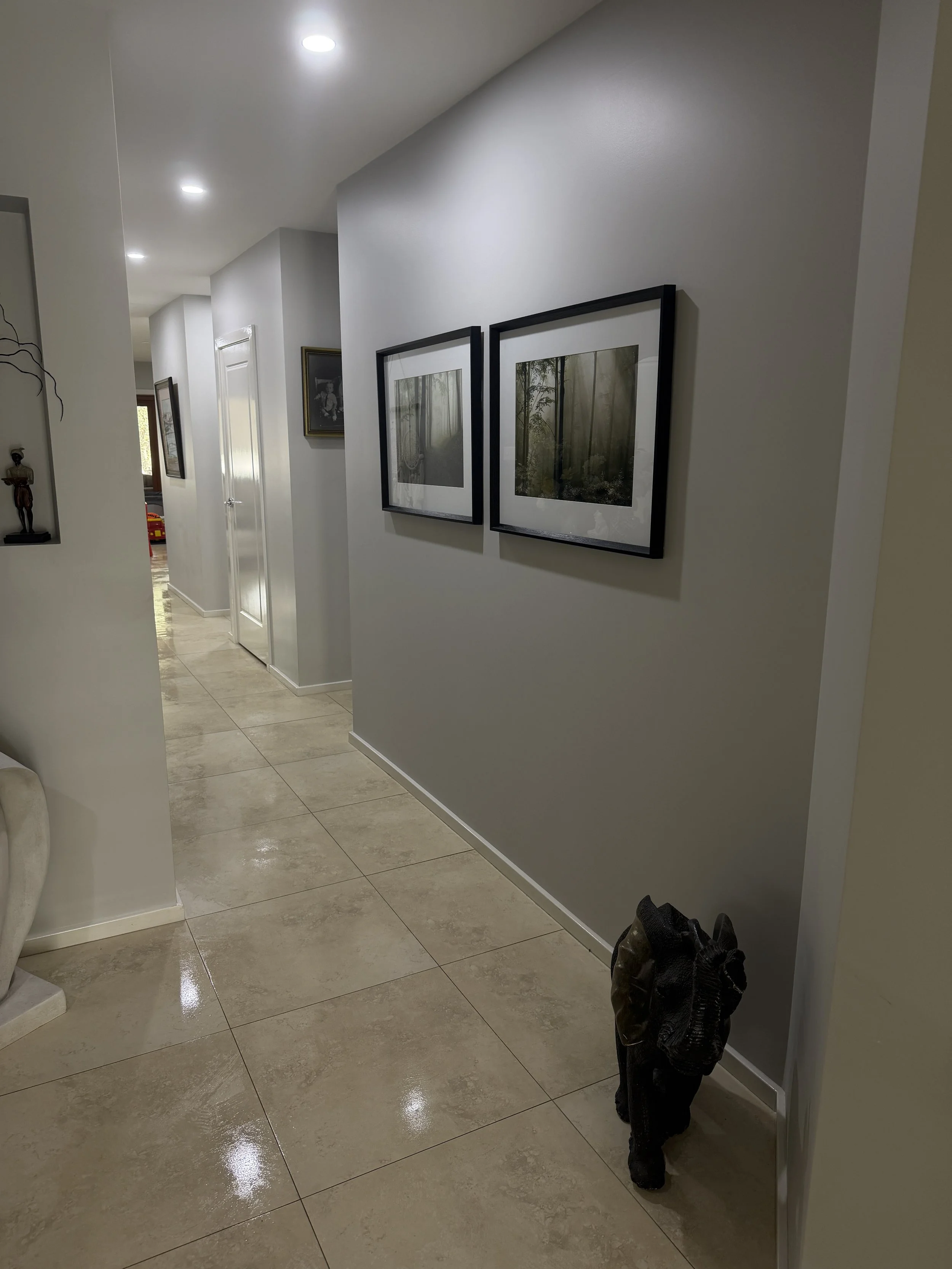 A hallway with beige tiled floors, gray walls, and ceiling lights. There is a small sculpture on a wall shelf to the left and two framed pictures on the gray wall on the right. Near the bottom right, a pair of shoes is visible.