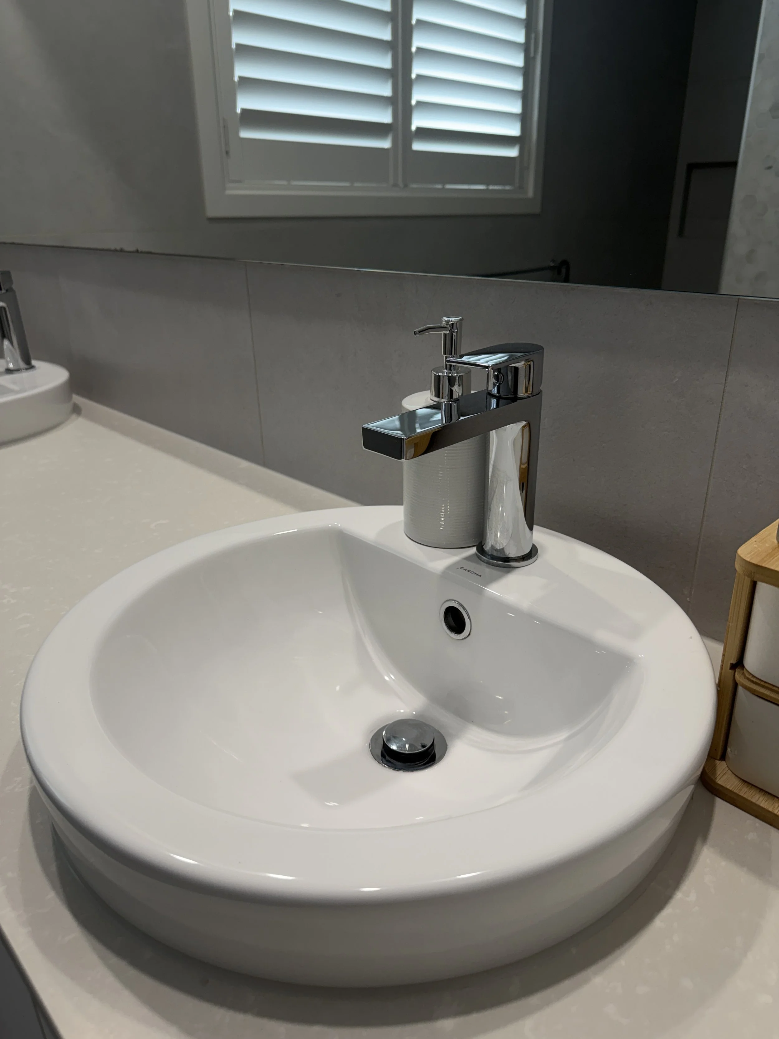 Close-up of a white bathroom sink with a modern chrome faucet, a mirror above, and a window with white shutters in the background.