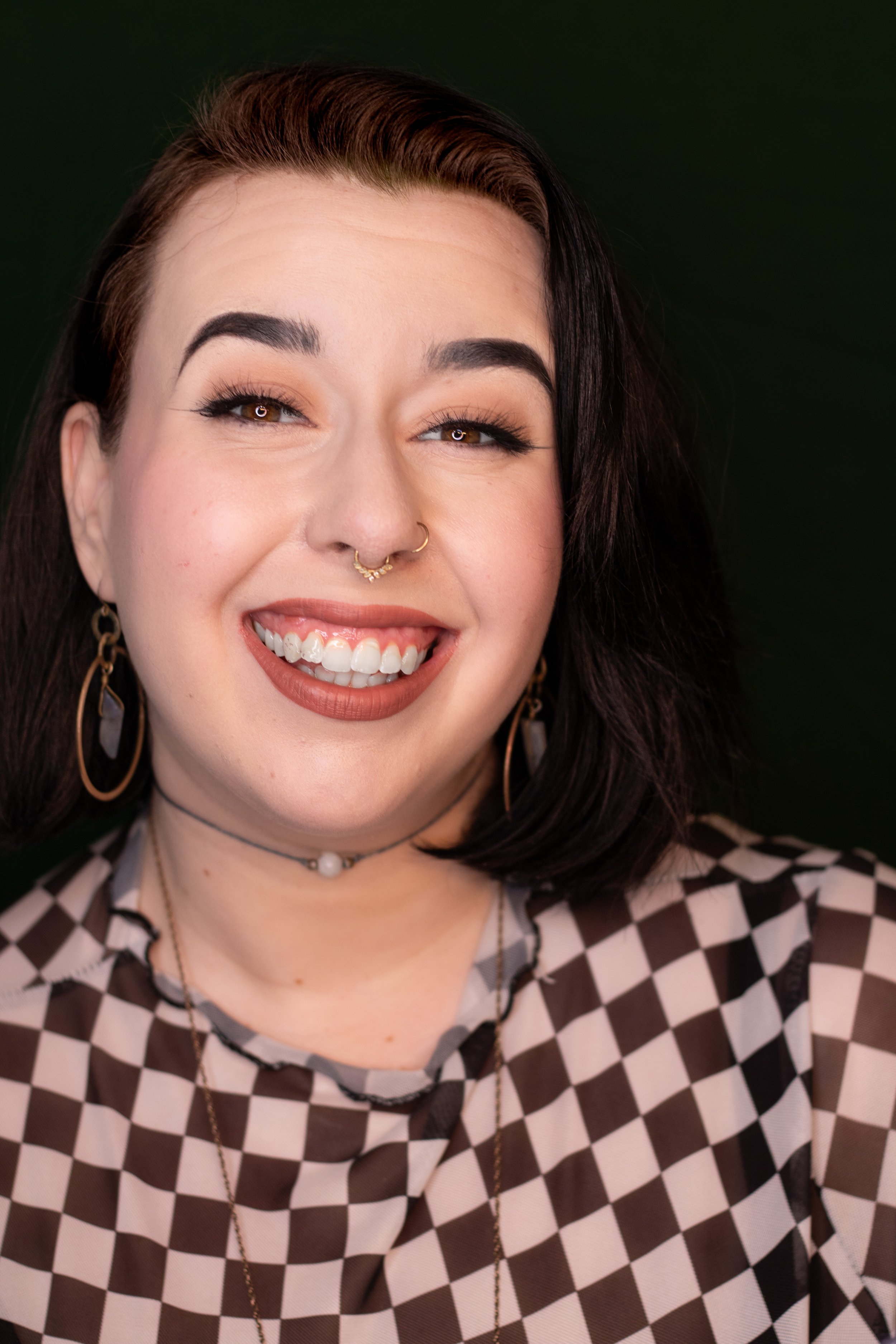 A smiling woman with brown hair, light skin, and various facial piercings including a septum ring and nose ring, wearing earrings, necklaces, and a black-and-white checkered top, against a dark green background.