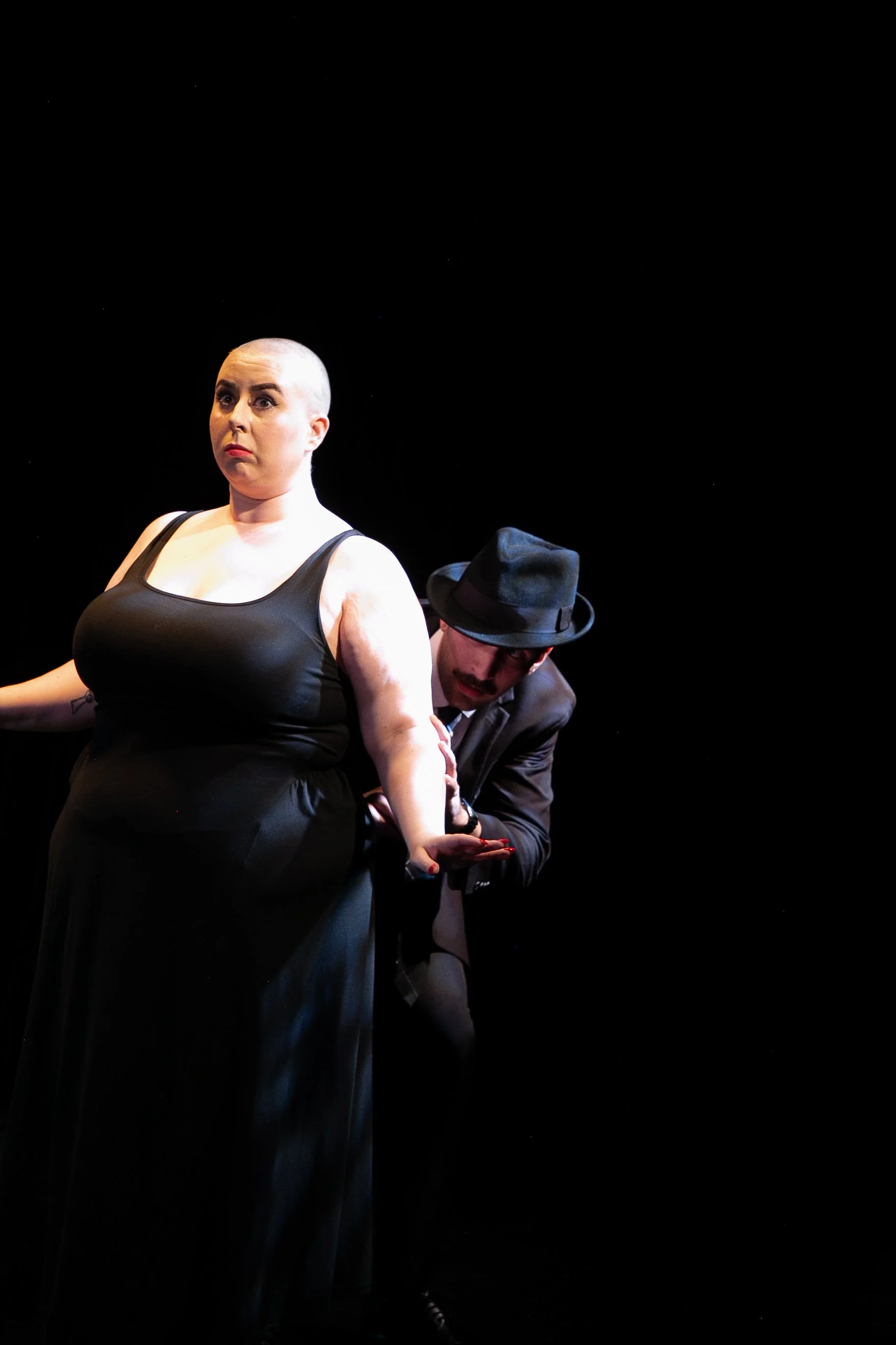 A woman with a shaved head wearing a black dress, standing on stage with a serious expression, and a man wearing a black hat and suit behind her.