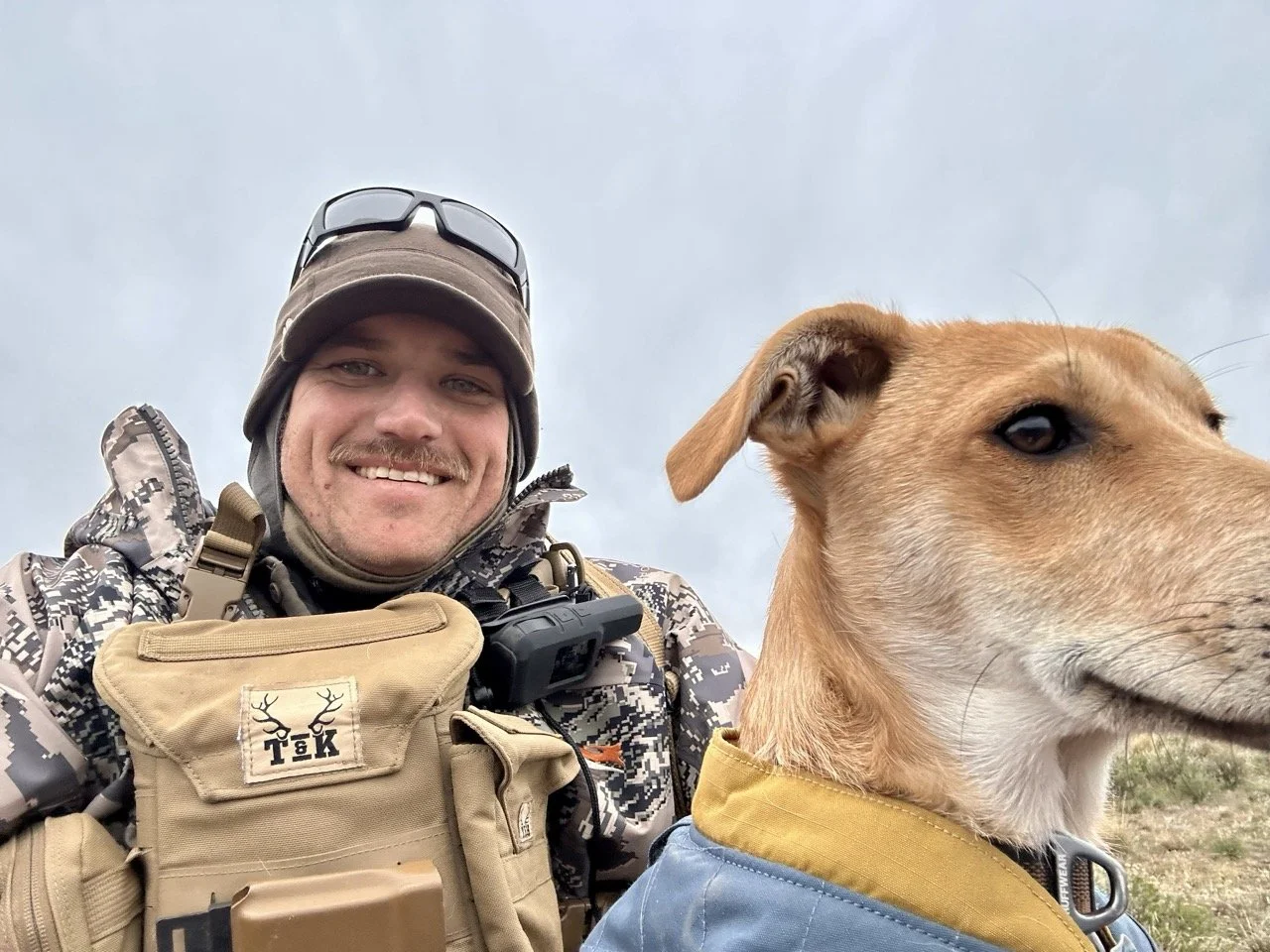 Close-up of a smiling man in camouflage outdoor gear with a tan tactical pouch and sunglasses on his head, posing outdoors with a tan dog in a blue jacket.