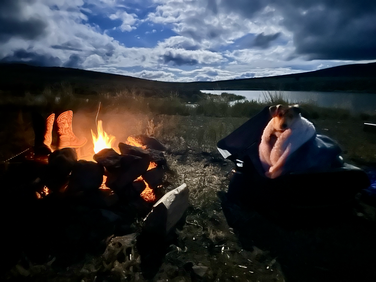 A nighttime outdoor scene near a body of water with a campfire, a pair of cowboy boots on the ground, and a dog in a blanket, looking at the camera.