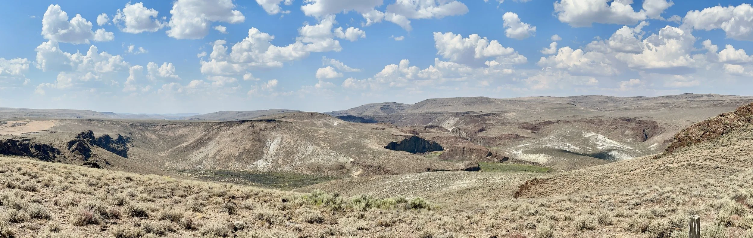 A dry, arid landscape with rolling hills and deep valleys, under a blue sky with scattered white clouds.
