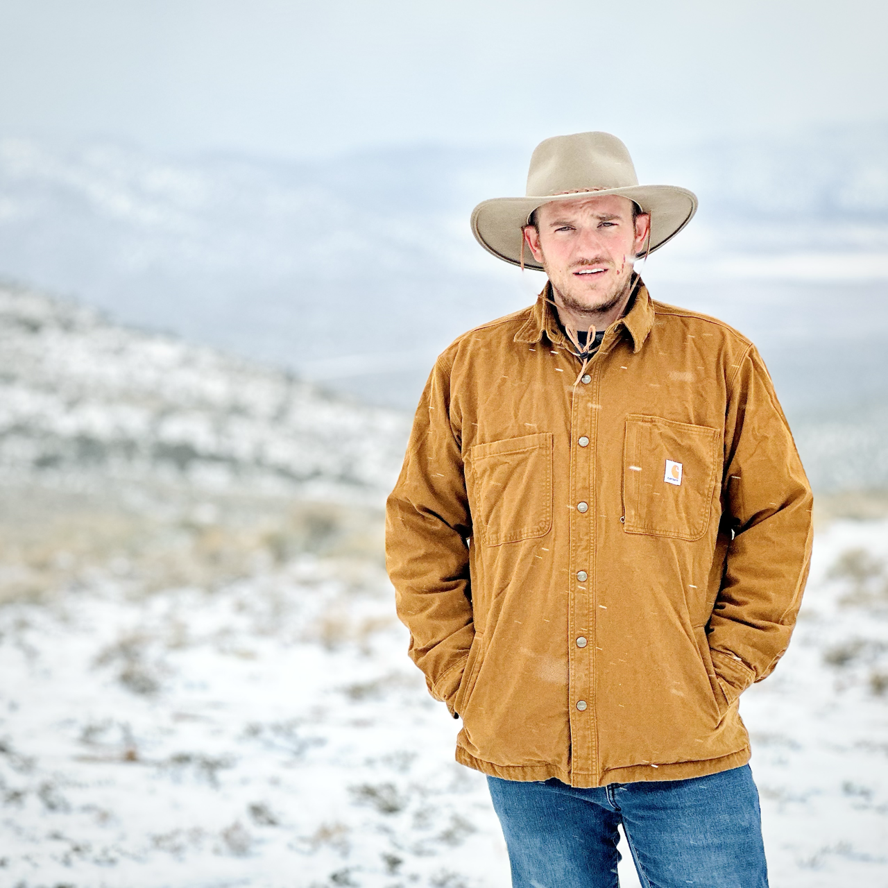 A man standing outdoors in a snowy landscape, wearing a brown Carhartt jacket, blue jeans, and a wide-brimmed beige hat, with hands in pockets.