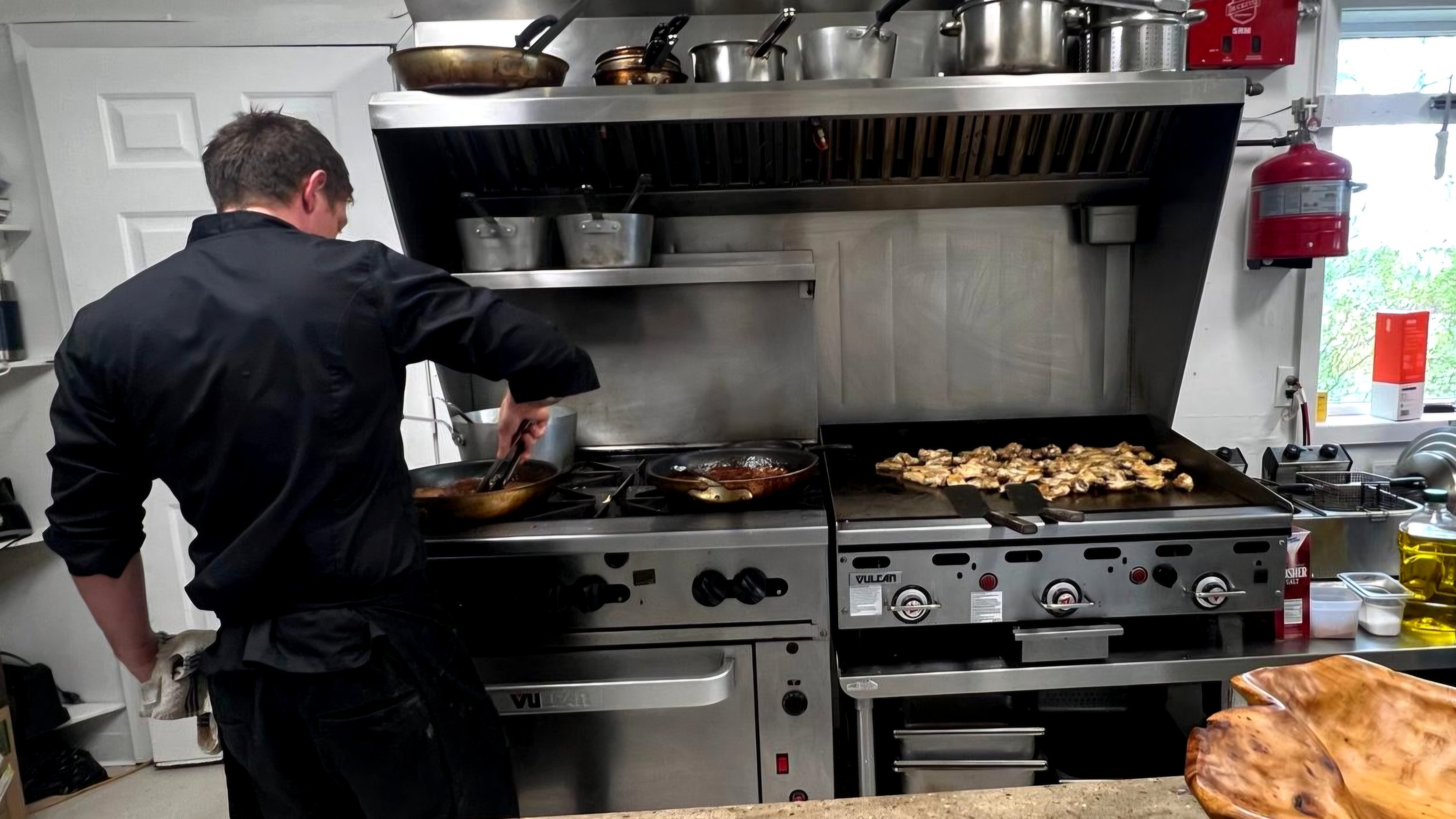 A chef cooking on a commercial stove with various pots and pans, including grilled chicken on the flat-top grill, in a professional kitchen. Fire extinguisher, cooking ingredients, and kitchen utensils are also visible.