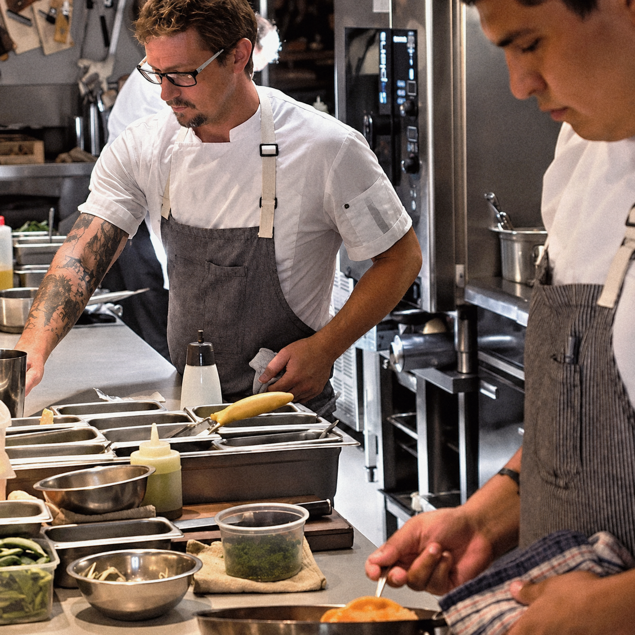 Two chefs in a professional kitchen preparing food, with various ingredients and kitchen tools visible on the counter.
