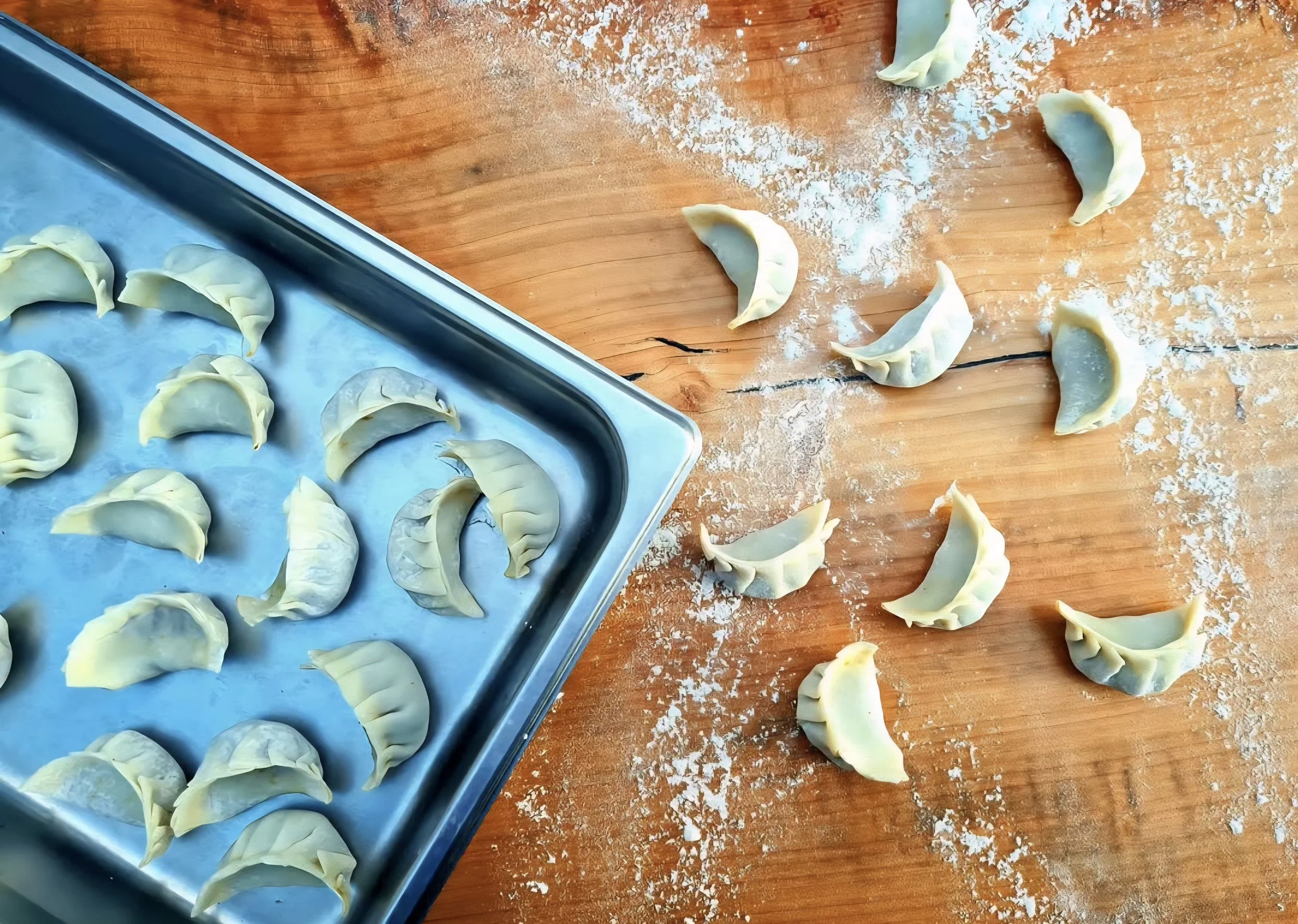 A baking sheet with uncooked dumplings on parchment paper, sprinkled with flour, on a wooden surface with scattered dumplings.