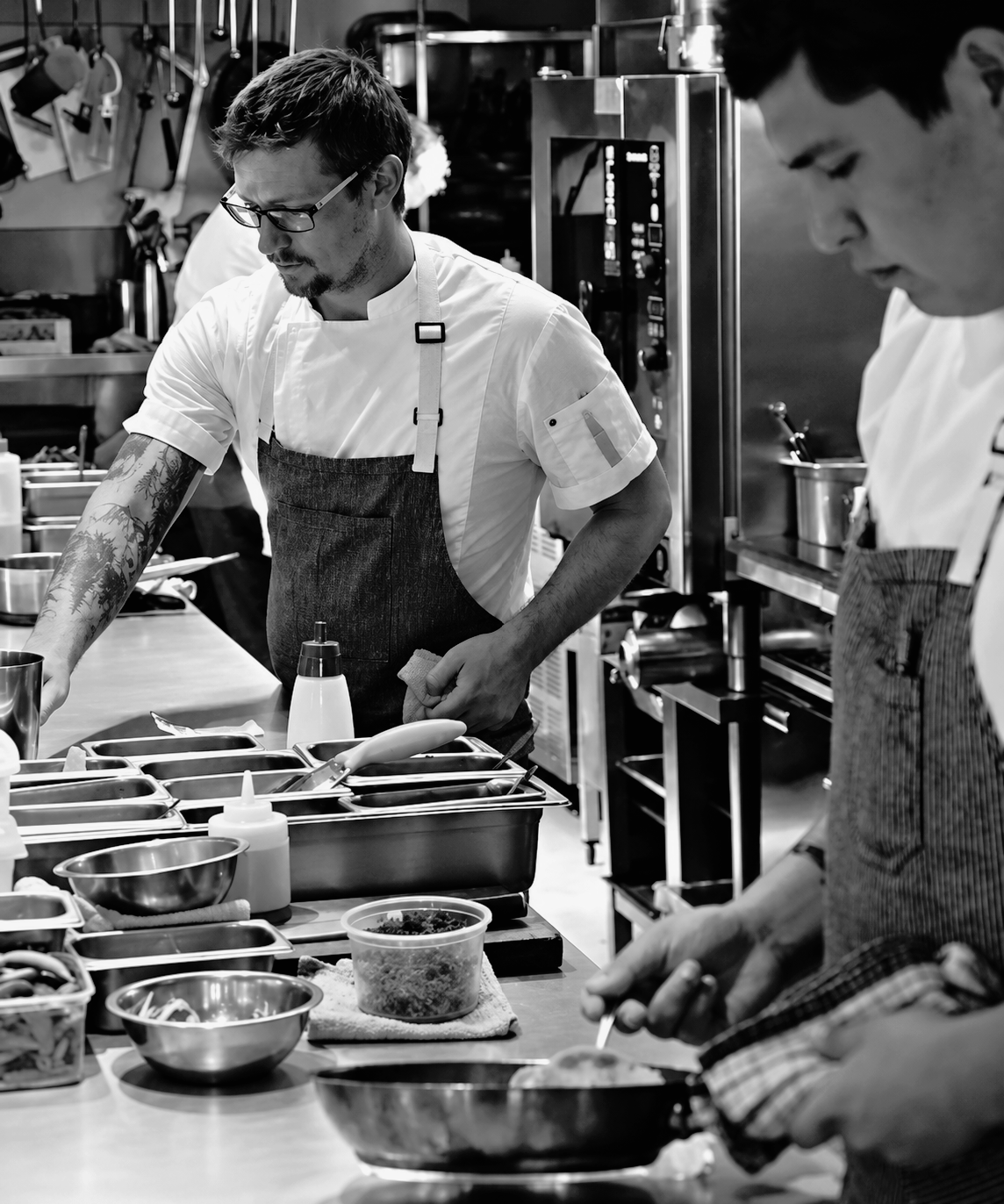 Two chefs working in a professional kitchen, preparing food at a countertop with various utensils and ingredients.