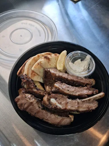 A black takeout container with short ribs, grilled meat, and potatoes, alongside a small container of sauce, placed on a metal surface with a clear plastic lid nearby.
