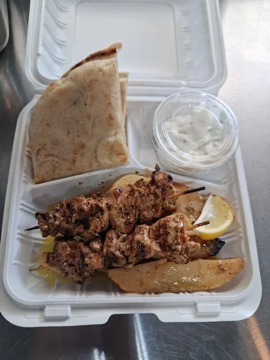 A takeout meal in a white foam container with a flatbread, grilled meat skewers, potato wedge, lemon wedge, a container of creamy white sauce, and a folded flatbread.