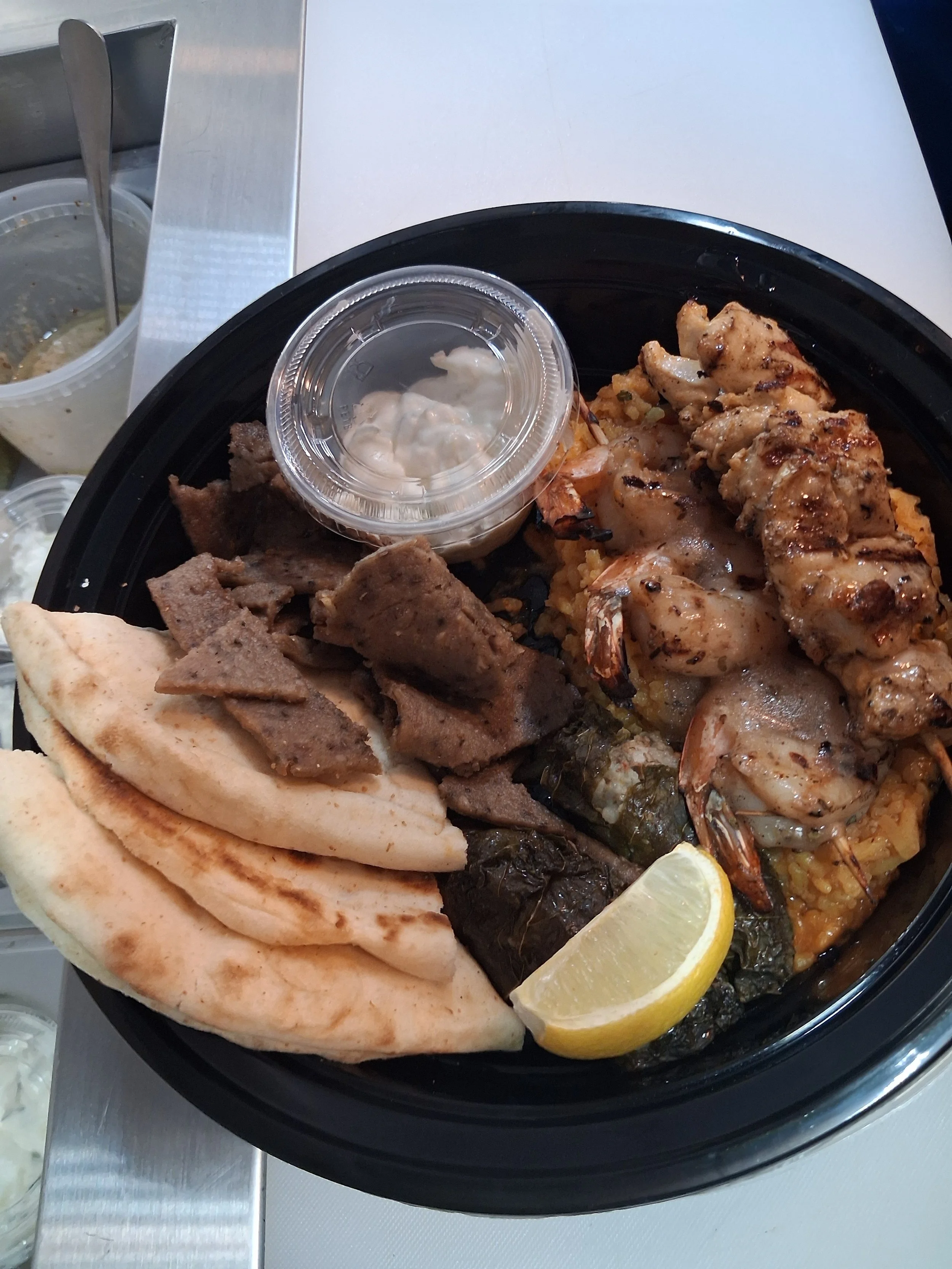 A black plate with grilled chicken pieces, shrimp, rice, a lemon wedge, and a small container of sauce, with pita bread and condiments on the side.