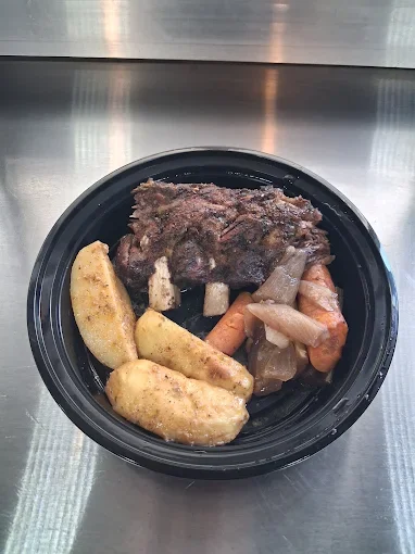 Cooked beef with potato wedges, carrots, and celery in a black plastic tray on a metal surface.