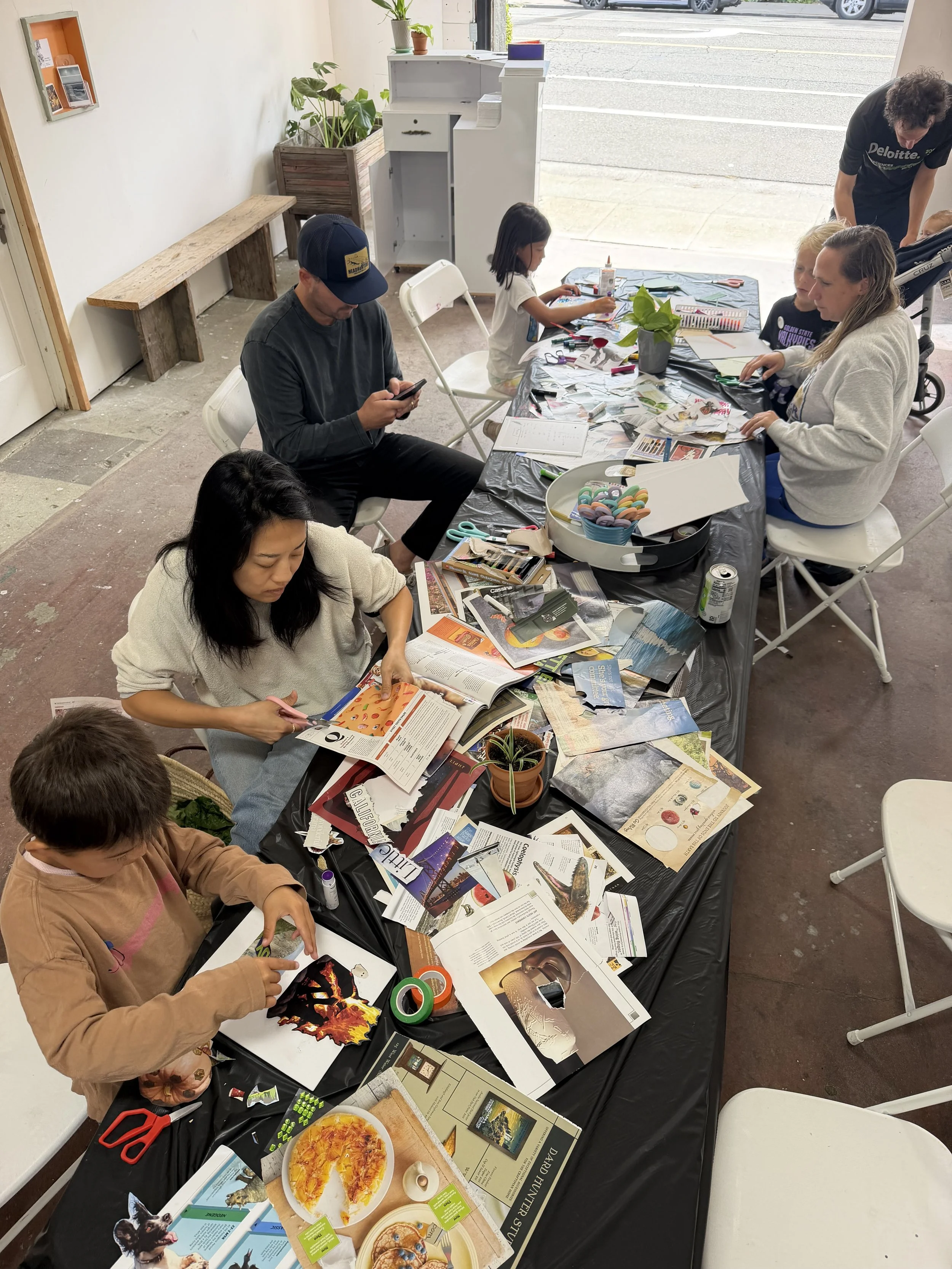People sitting at a long table engaging in arts and crafts activities. The table is covered with magazines, papers, scissors, and other craft supplies. Inside, there are children and adults, some cutting paper, while others are working on projects. Outside the open door, parked cars are visible. The room has a rustic floor, a bench against the wall, and some potted plants.