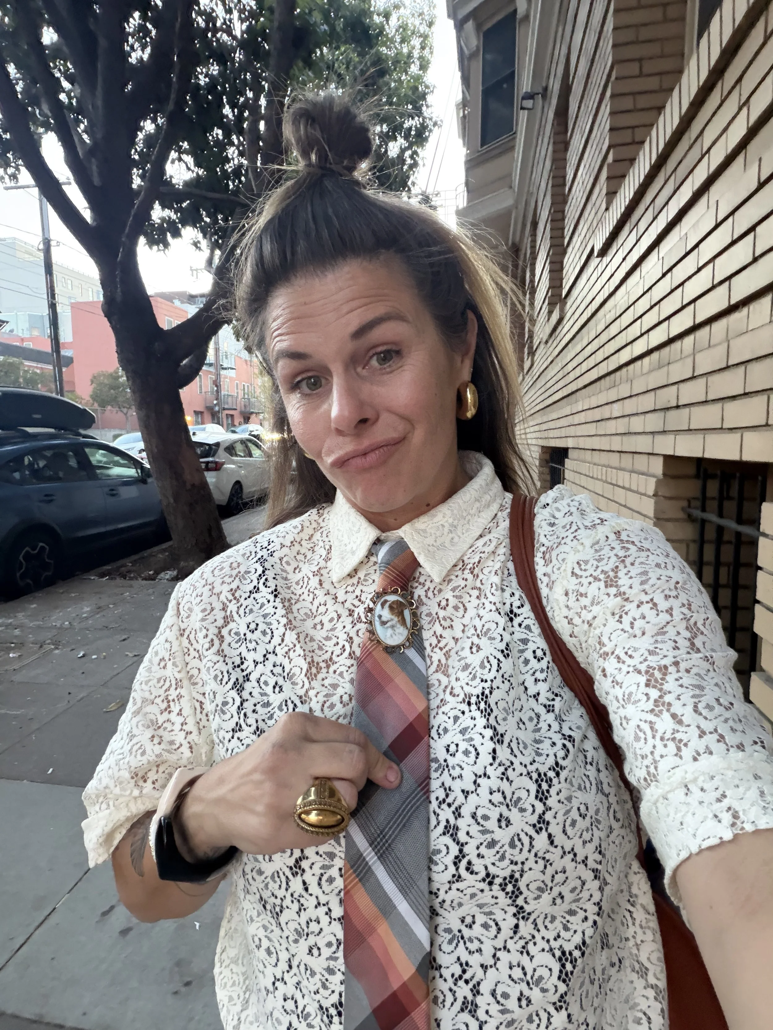 A woman wearing a beige lace top, plaid tie, and gold jewelry, holding a plaid tie in front of a brick building, with cars parked on the street and a tree in the background.
