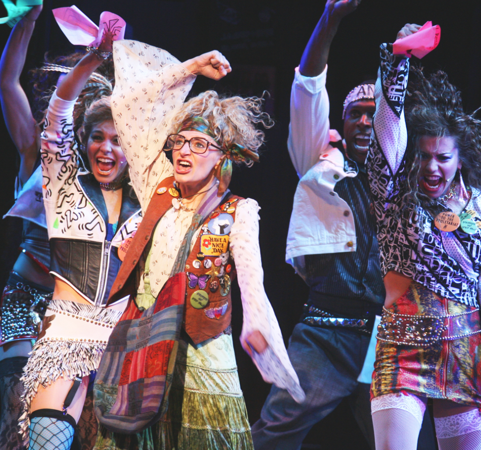 Performers on stage dressed in colorful, eclectic costumes, expressing energetic dance moves and vibrant facial expressions.