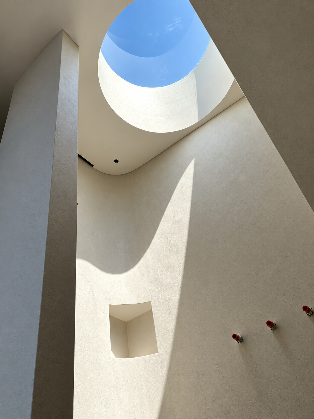 Interior view of a modern building with white textured walls, a circular skylight revealing a blue sky, and sharp shadows cast by the sunlight.