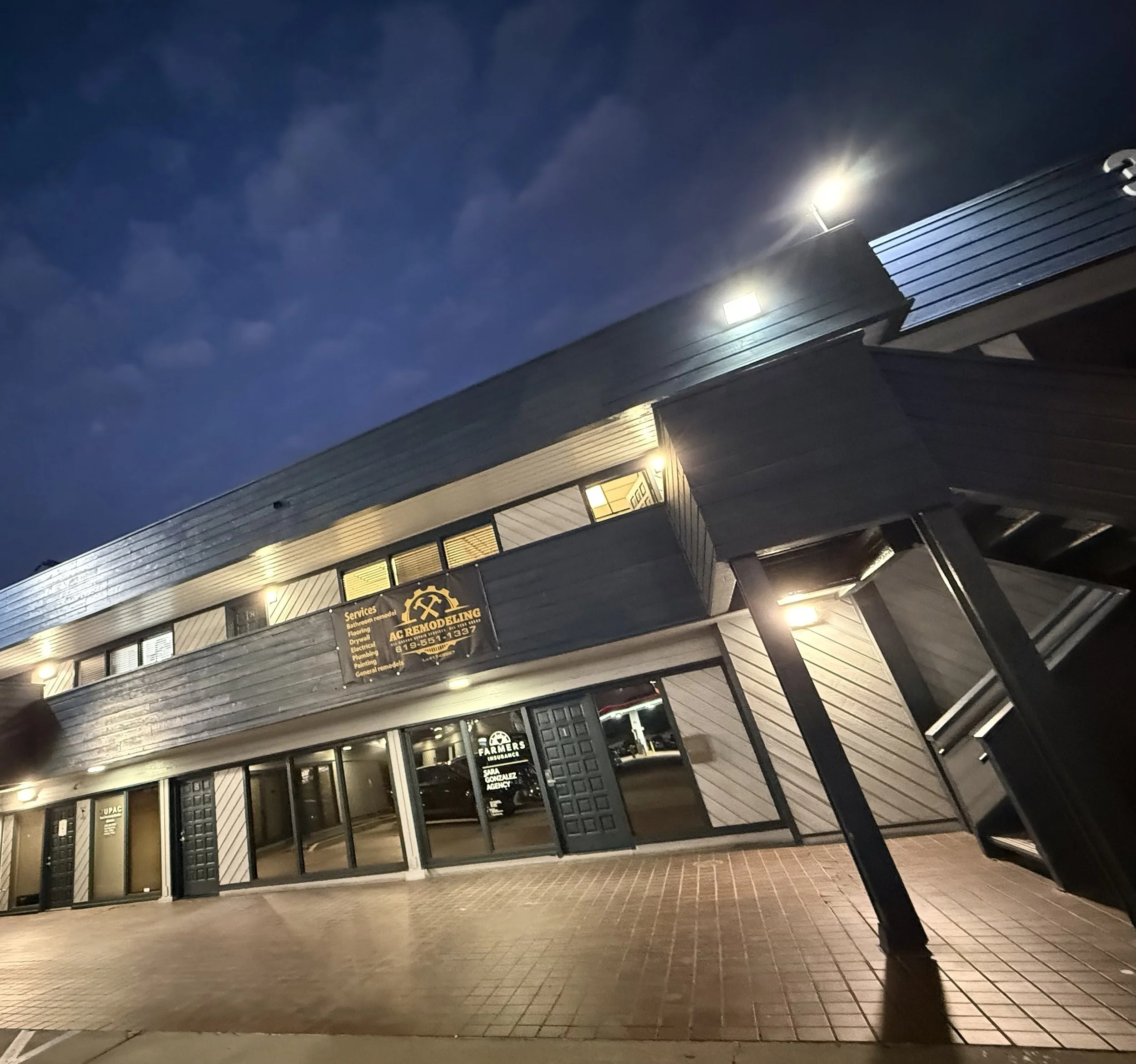 A modern commercial building with a sign for Acme Remodeling, illuminated at night, with a sidewalk in front and a clear dark blue sky above.