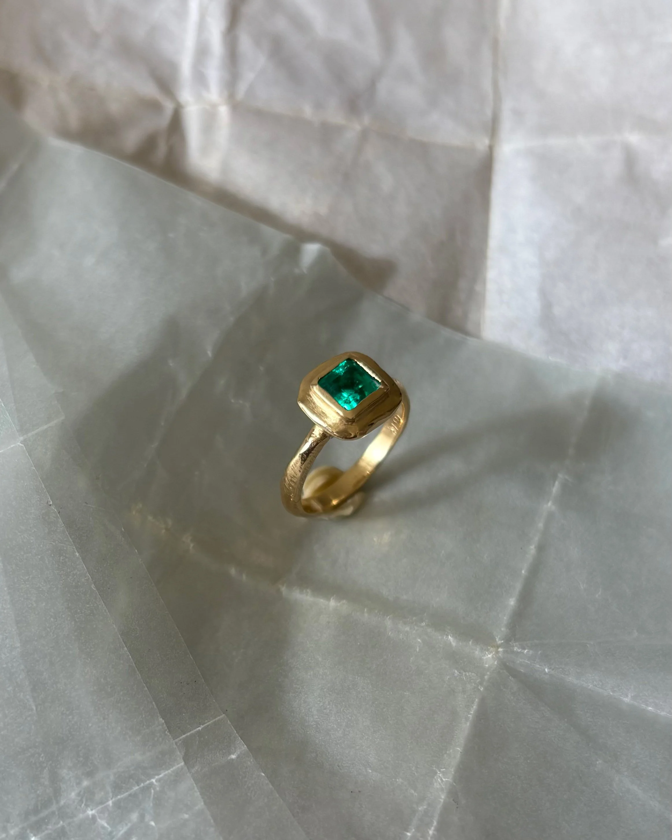 A gold ring with a square emerald gemstone on a grayish surface, with crumpled white tissue paper in the background.