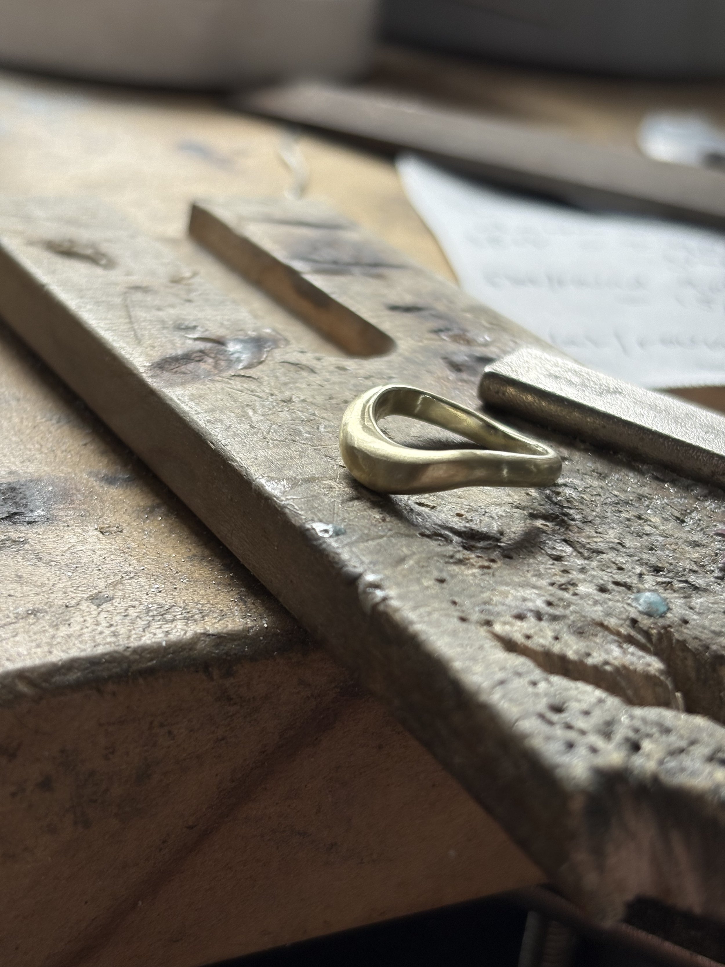 Close-up of a metal object, possibly a jewelry piece or a small component, resting on a textured, aged surface with tools and papers in the background, indicating a craft or workshop setting.