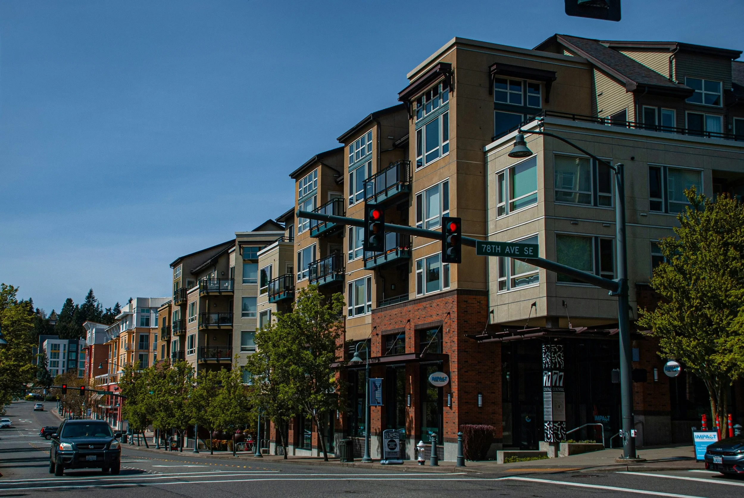 A city intersection with a building featuring multiple balconies and large windows, street signs indicating 78th Ave SE, cars on the road, traffic lights, and street trees under a clear blue sky.