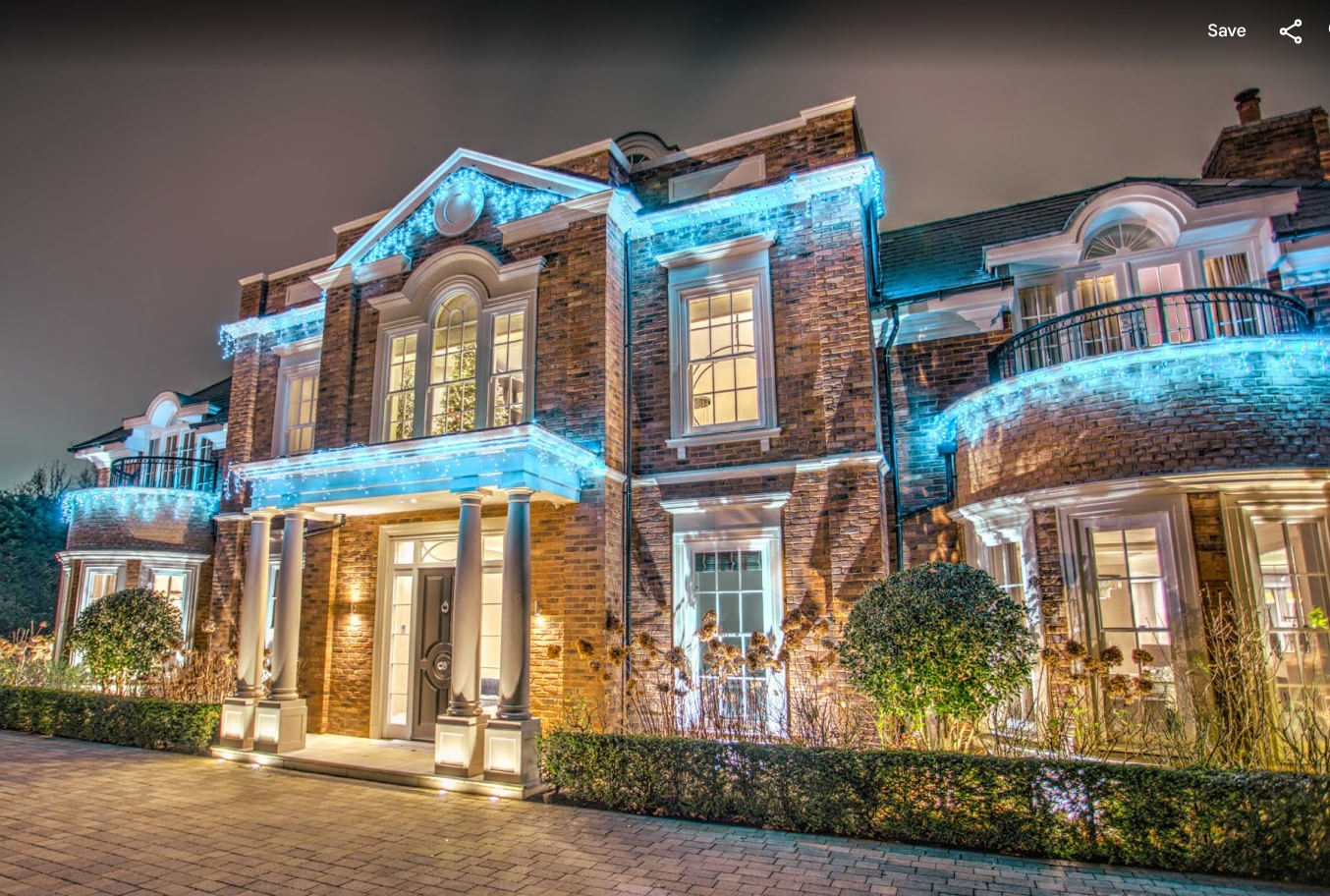 Nighttime view of a large brick house decorated with blue Christmas lights, illuminated windows, and landscaping in the front yard.
