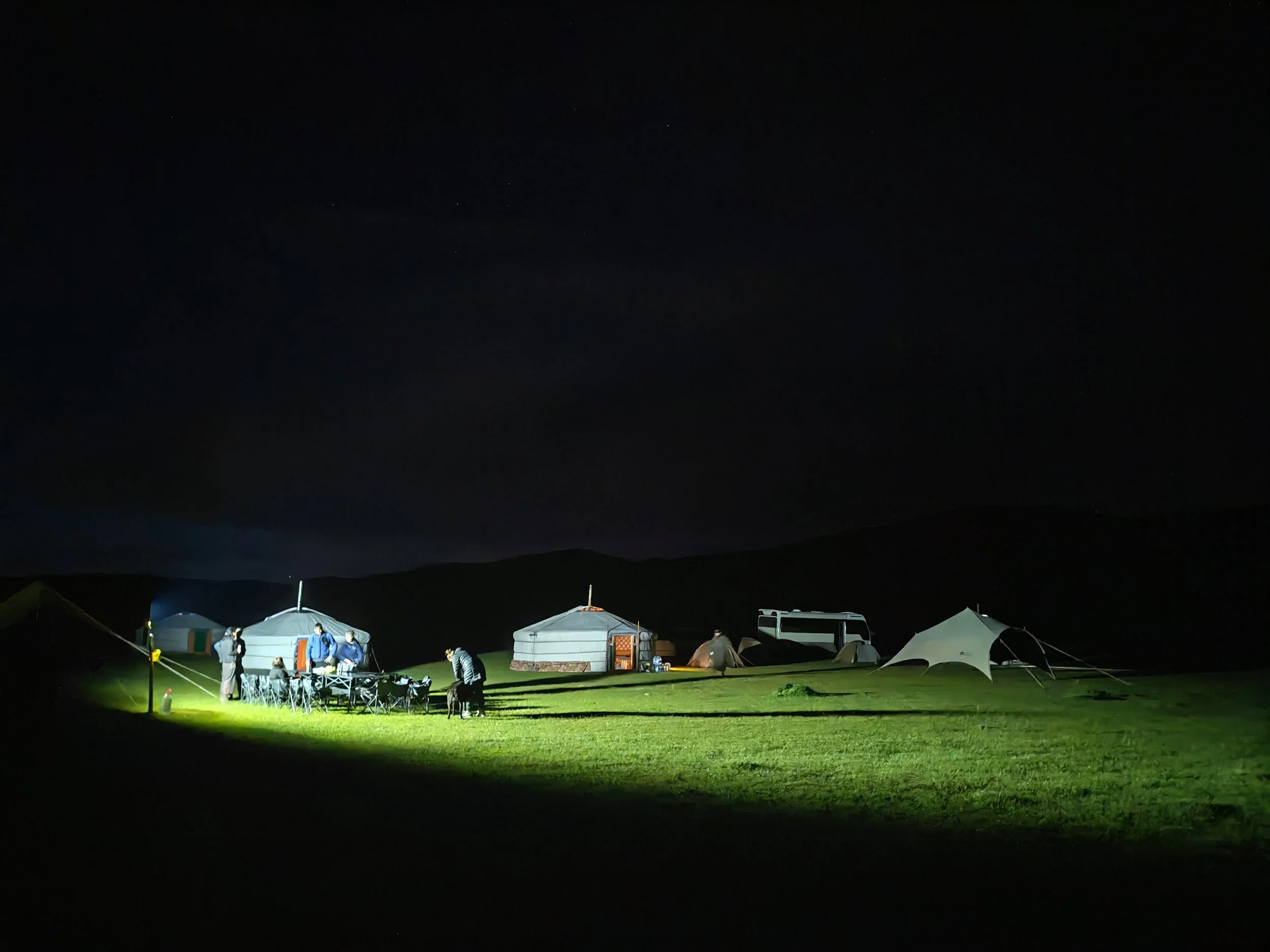 Night scene of a campsite with tents and people gathered around, illuminated by artificial lighting, under a dark sky with some stars visible.