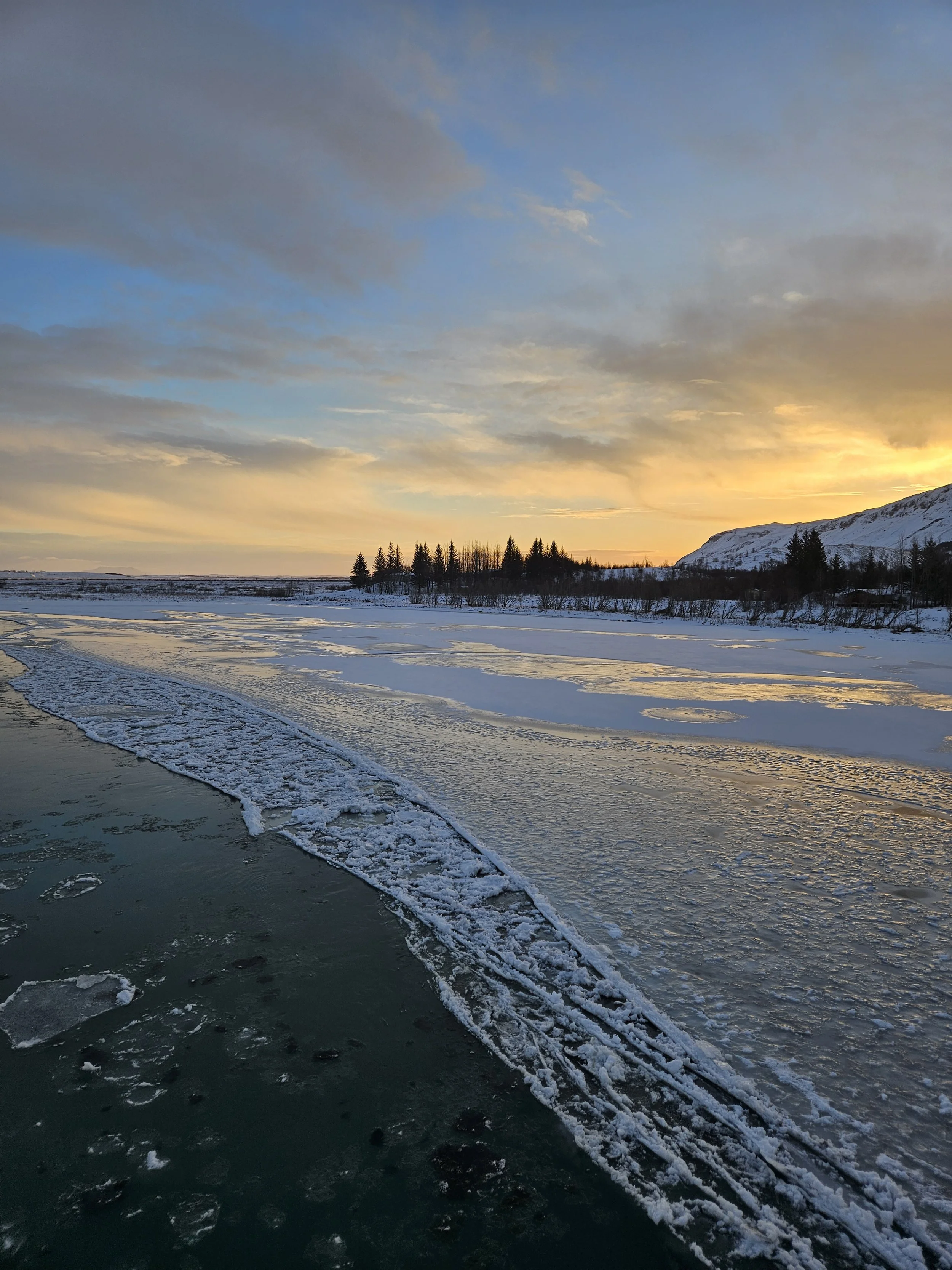 Frozen river with snow-covered banks, trees in the distance, mountains, and a sunset sky with clouds.