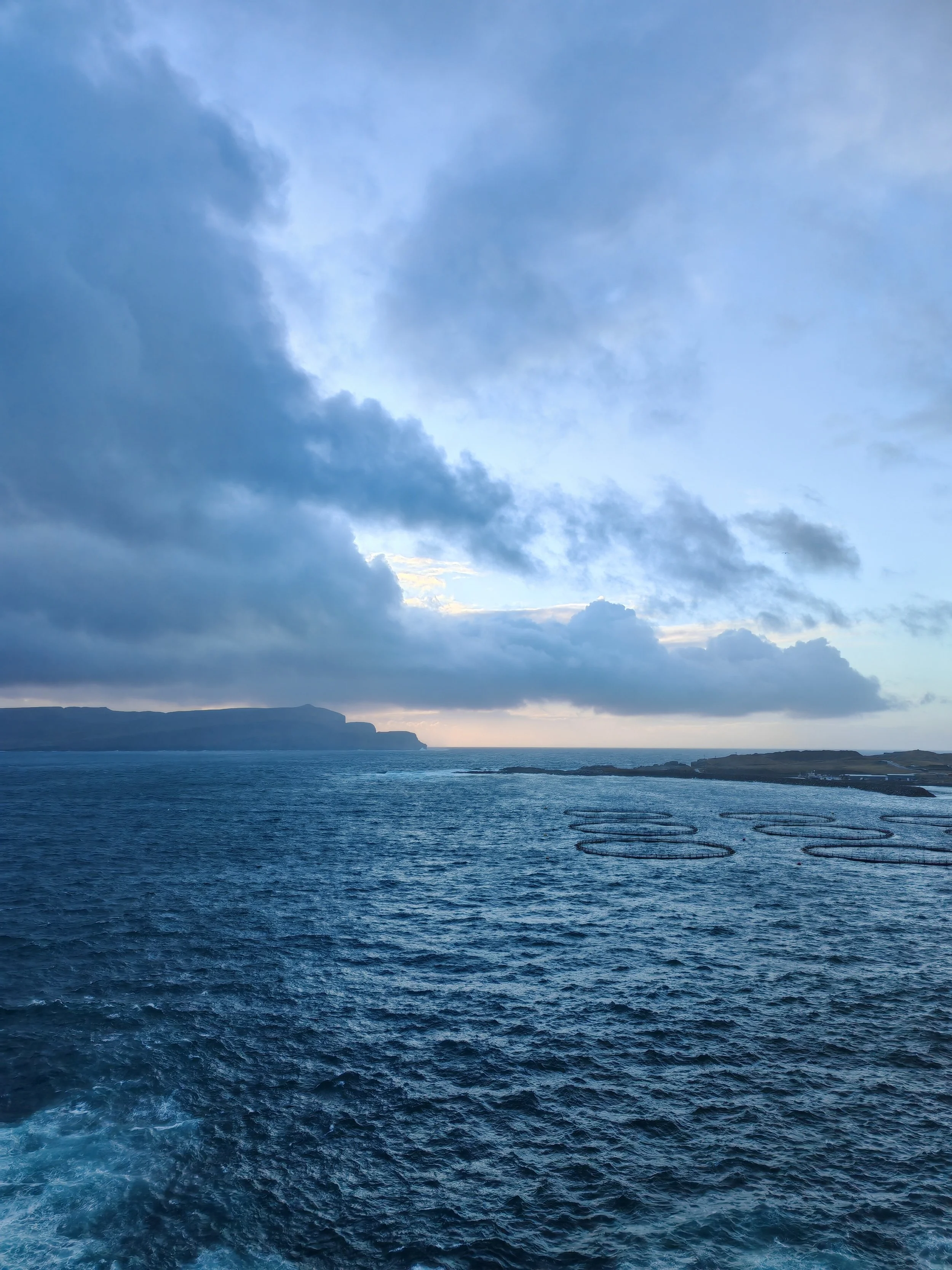 View of a deep blue ocean with visible wake pattern at the bottom, a distant landmass with cliffs on the horizon, and a sky filled with dark and light clouds.