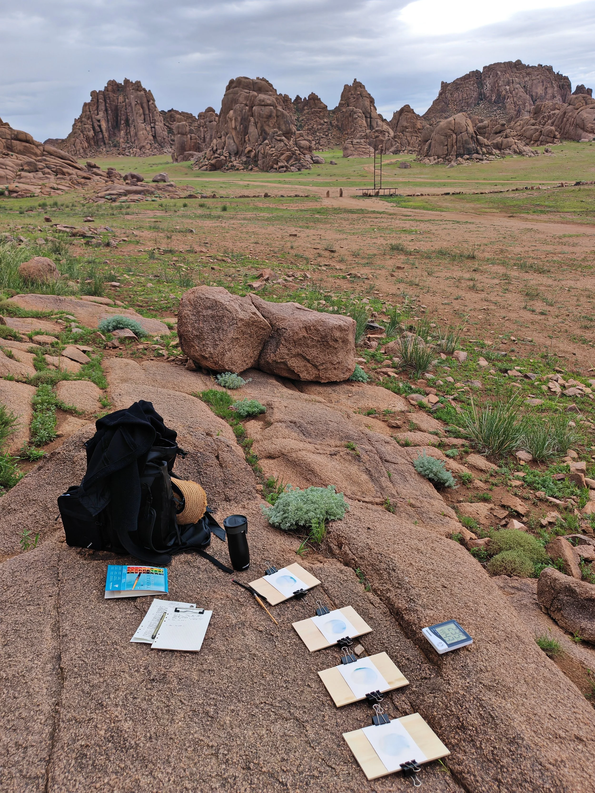 An outdoor scene in a rocky desert landscape with large rock formations and green patches, featuring scientific equipment on the ground including notebooks, a thermometer, and testing materials, with overcast skies.