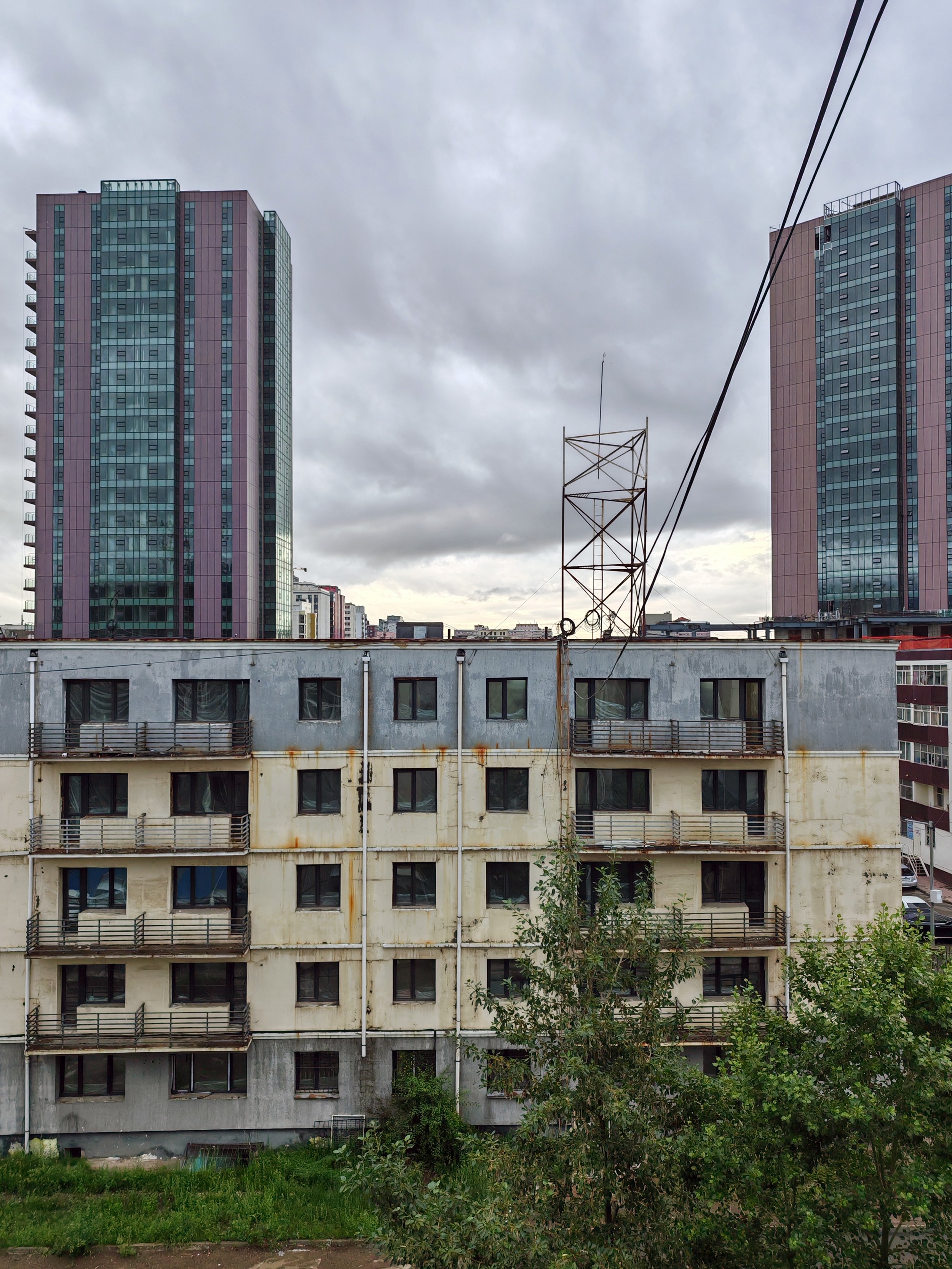 View of an urban landscape with tall modern glass buildings and an older, weathered building in the foreground, with some greenery at the bottom and a cloudy sky overhead.