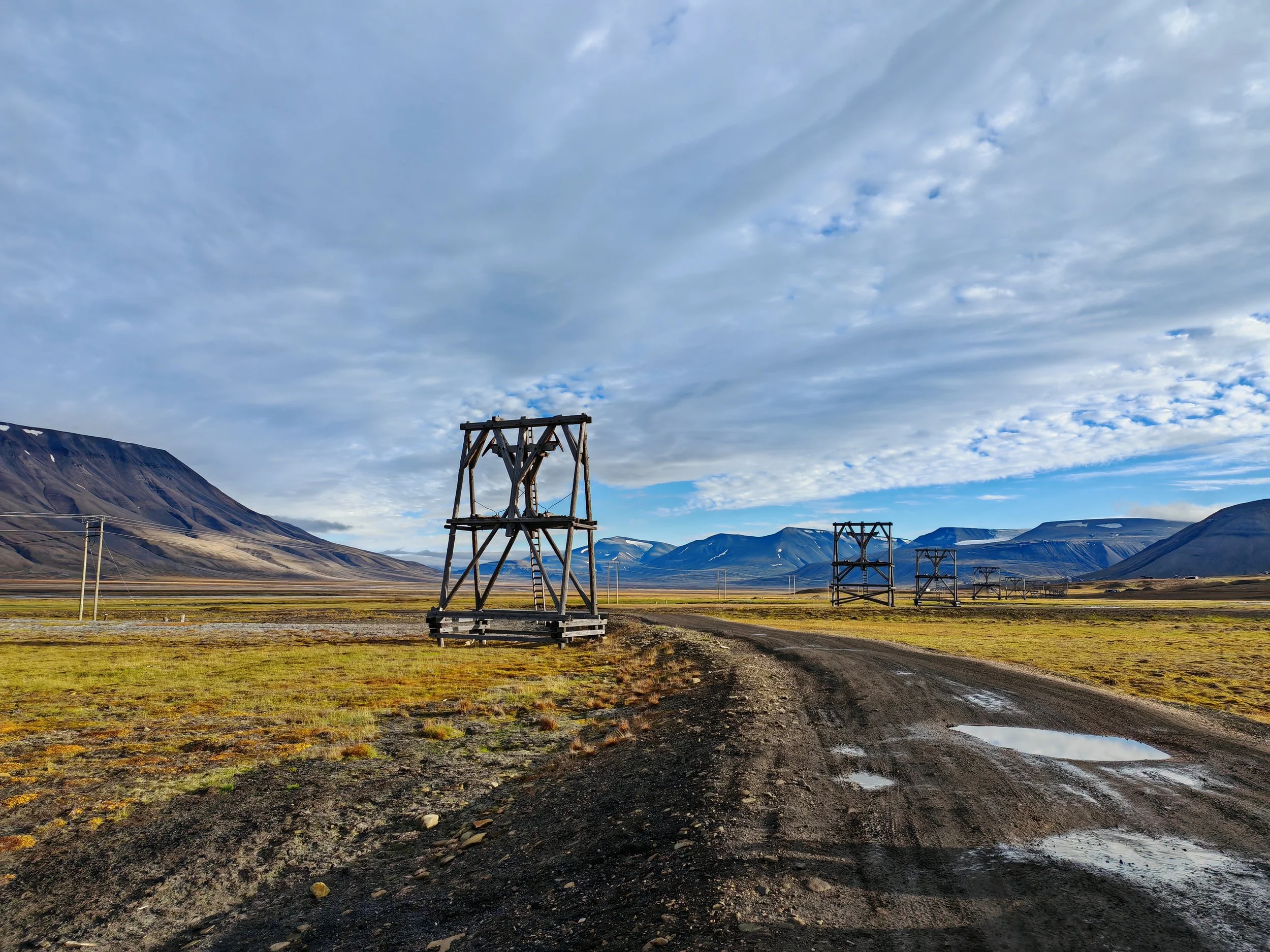 A landscape with mountains in the background, a dirt road in the foreground, and wooden power line towers scattered across a grassy field under a partly cloudy sky.