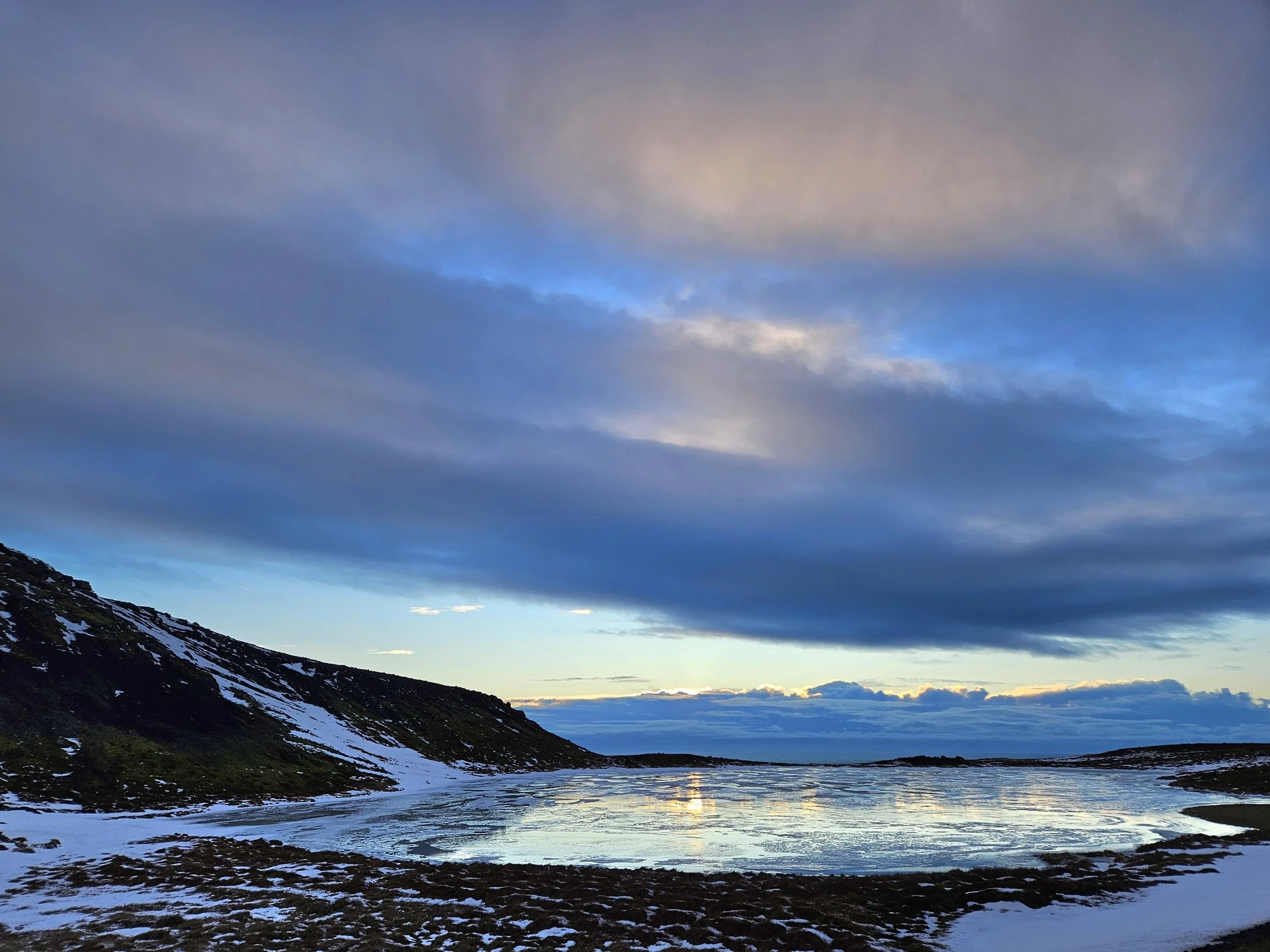 Scenic view of a mountain landscape with a partly cloudy sky, snow patches on the mountain slopes, and a body of water reflecting the sky.