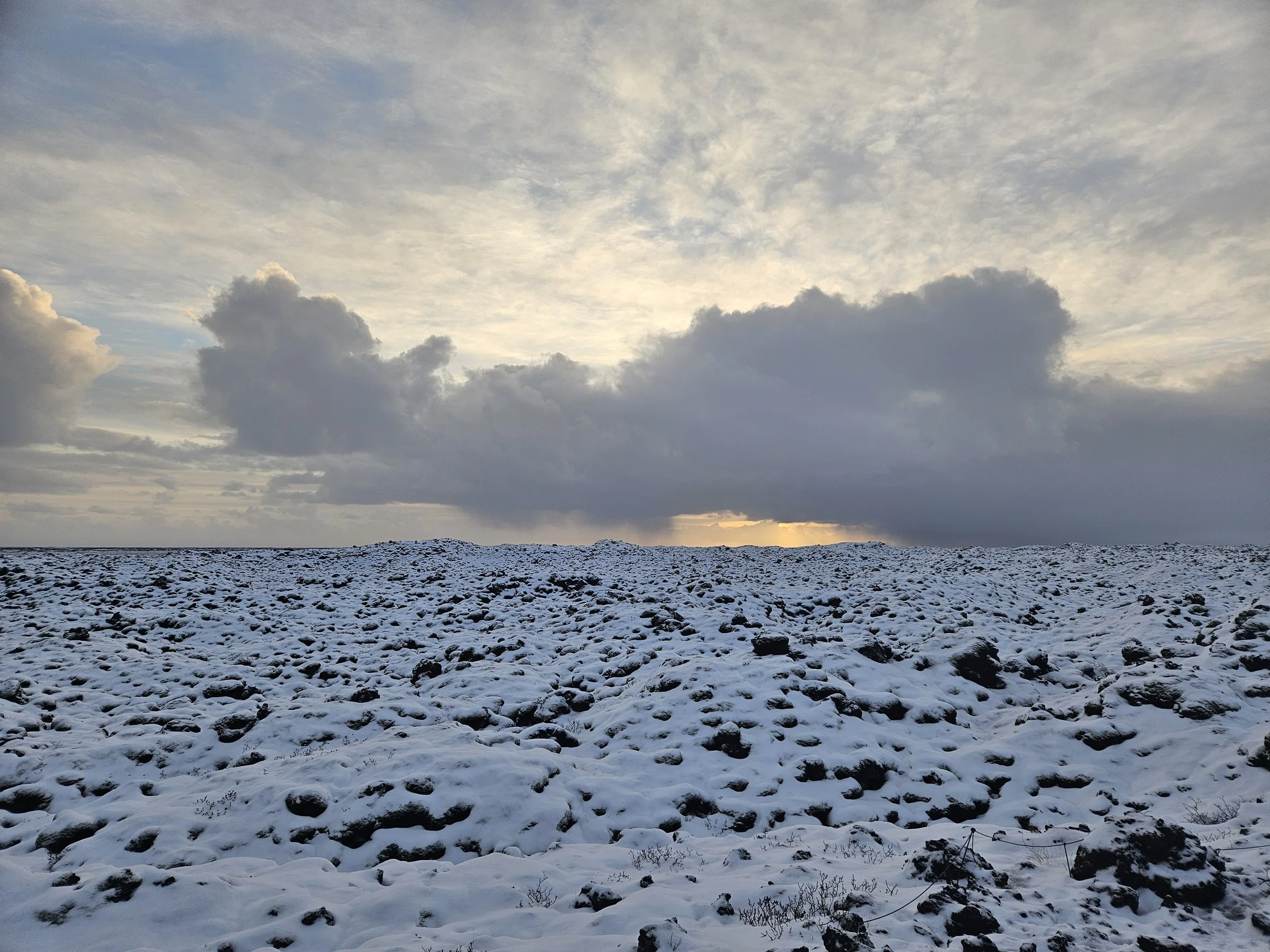 Snow-covered rocky terrain with a cloudy sky overcast, sunset or sunrise in the distance.