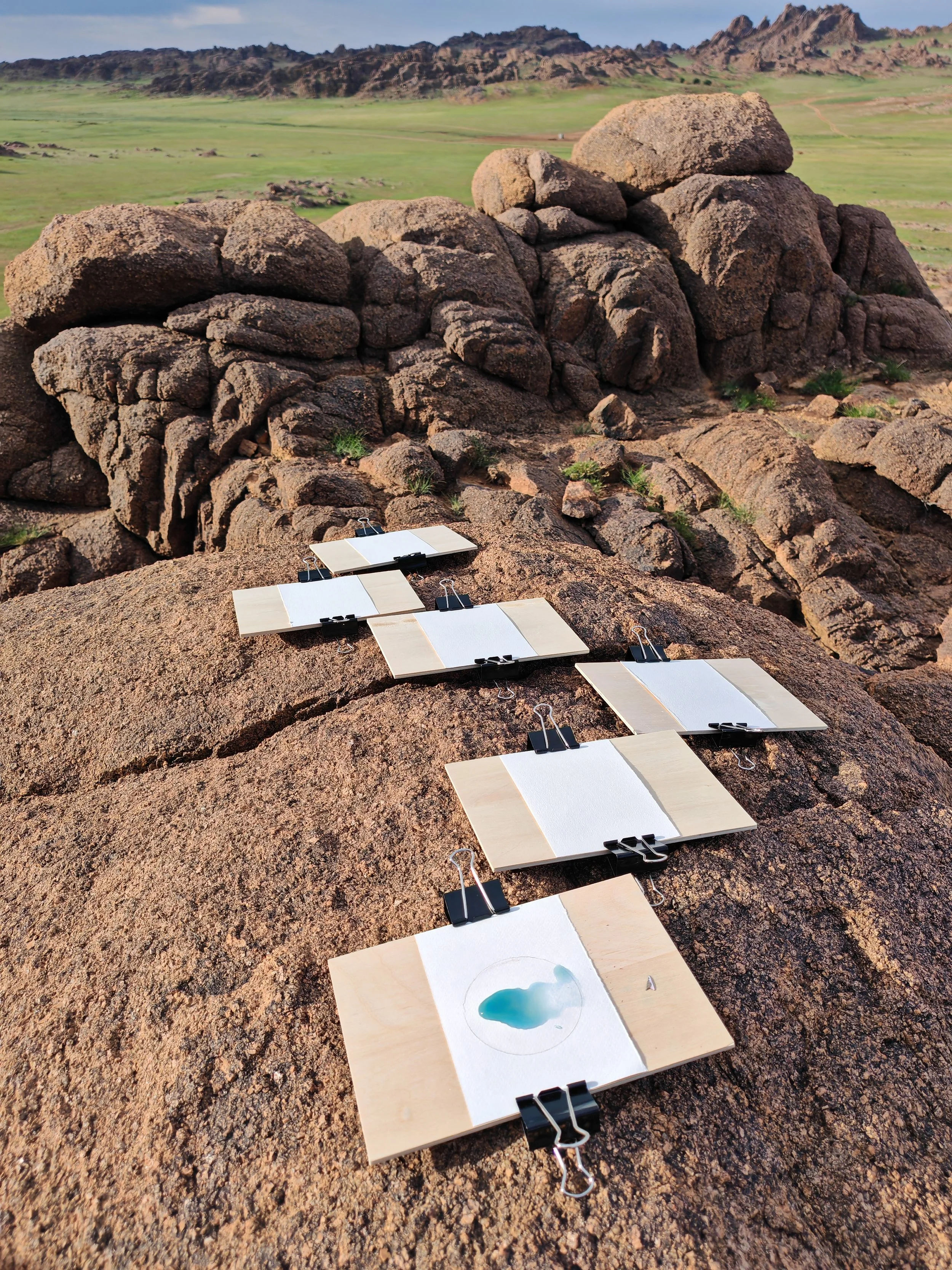Close-up of several watercolor paint samples on wooden boards, clipped with black binder clips, laid on rocky desert terrain with mountains and a green valley in the background.