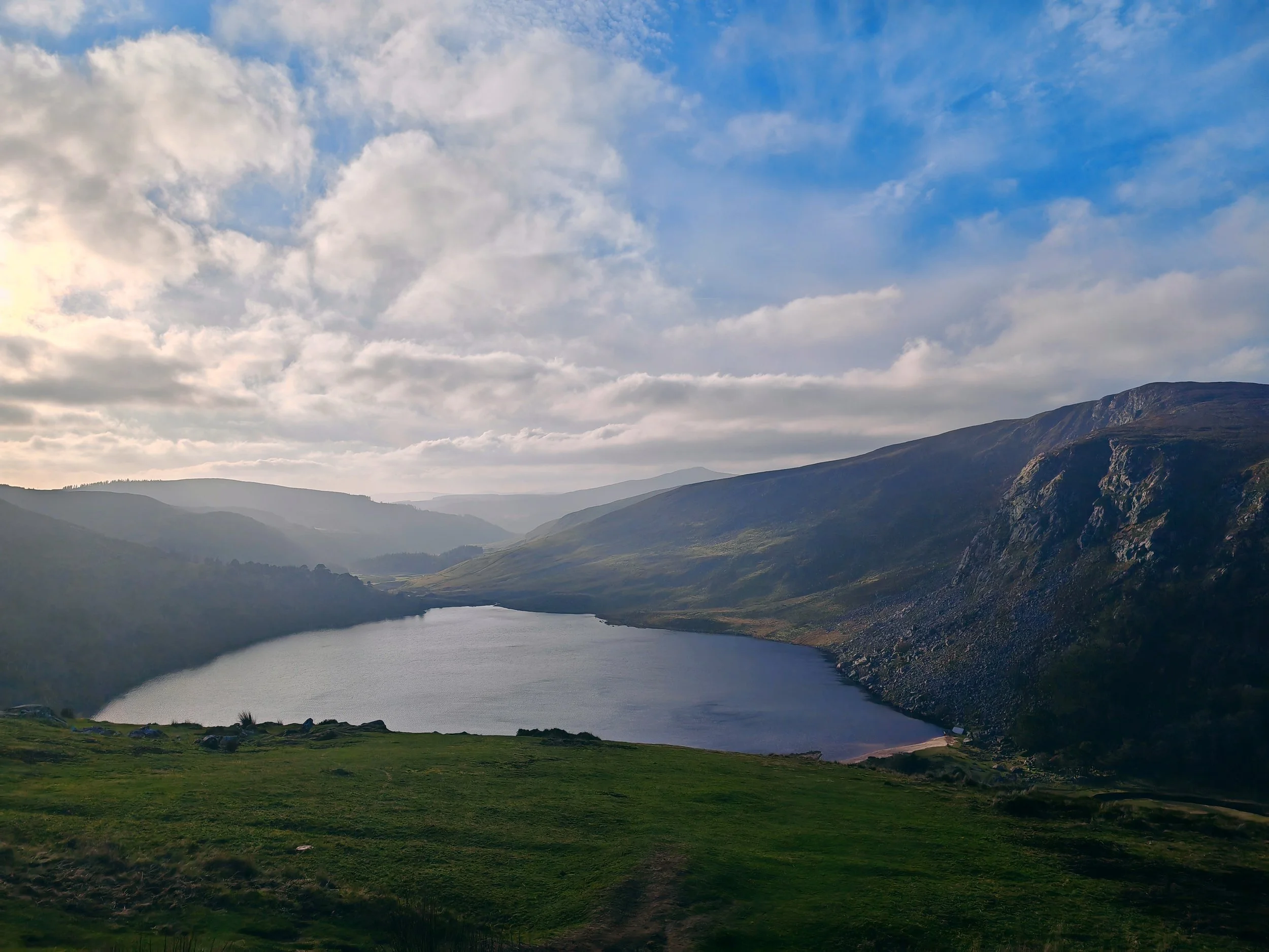 Scenic landscape with a river flowing through green grass and rolling hills, mountain range in the distance, and a partly cloudy sky above.
