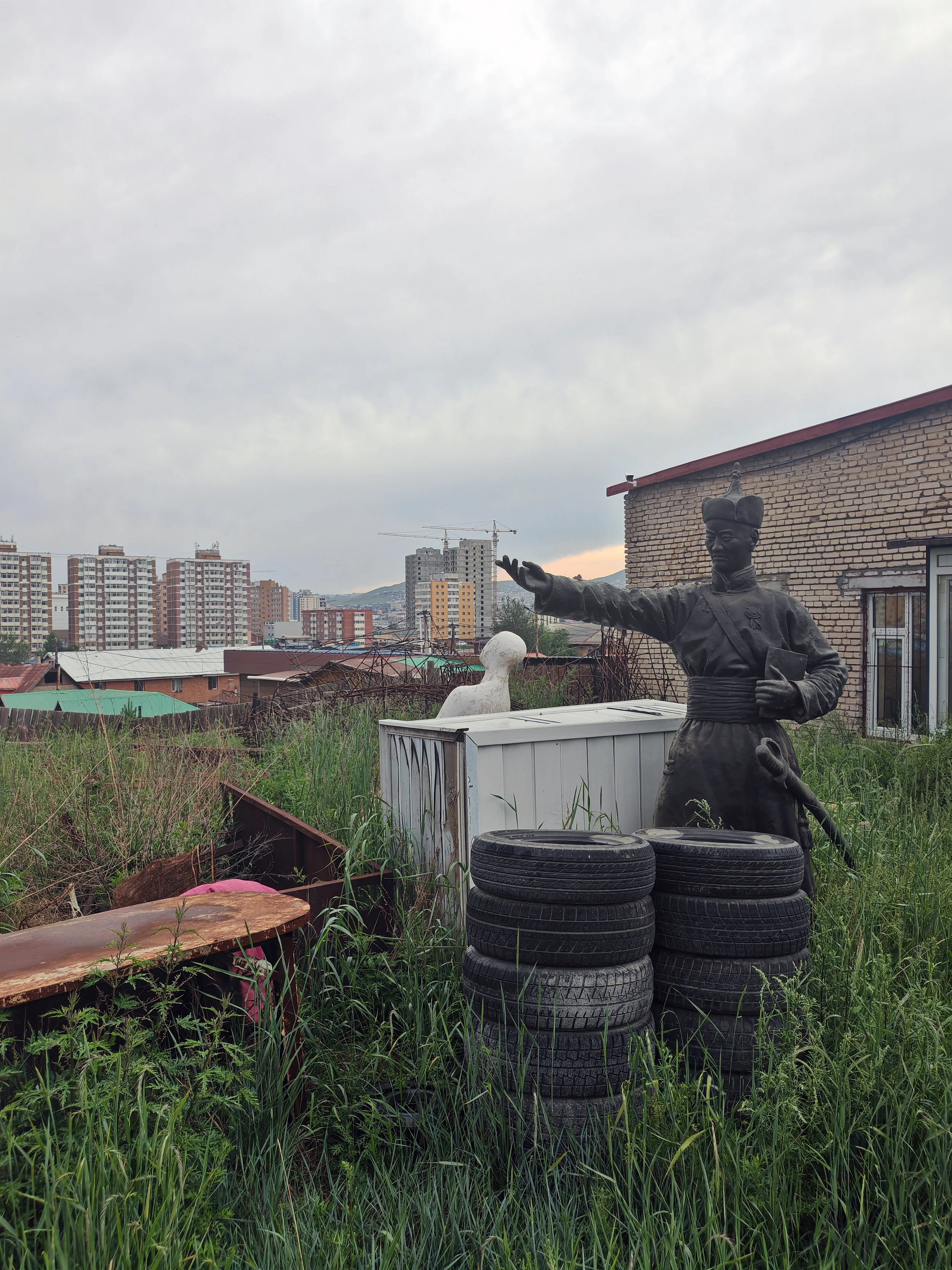 A cityscape with tall apartment buildings under a cloudy sky, featuring a bronze statue of a man in traditional attire extending his arm, surrounded by stacked tires, overgrown grass, and makeshift structures.